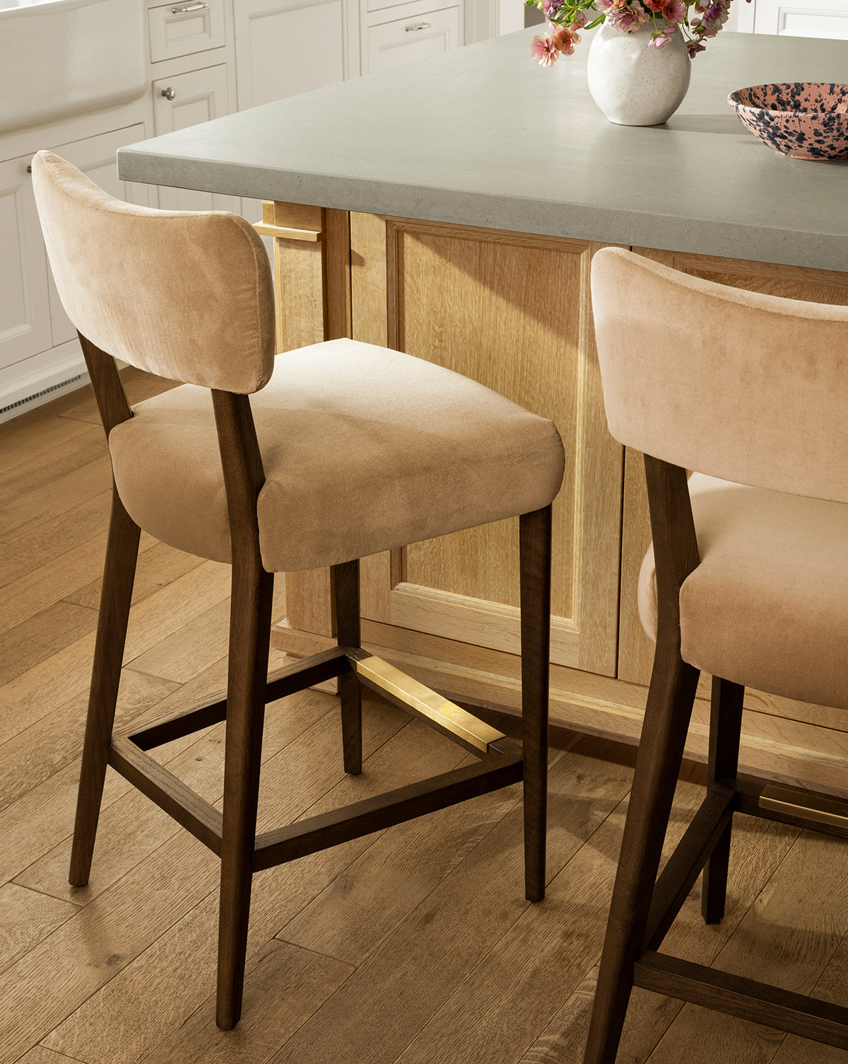 Two beige upholstered Makerspalm Etienne Stools (Ready to Ship) with dark wood legs sit by a light wood kitchen island with a gray countertop, where a vase and bowl accentuate the customizable stool design and complement the light wood floor. Vendixs
