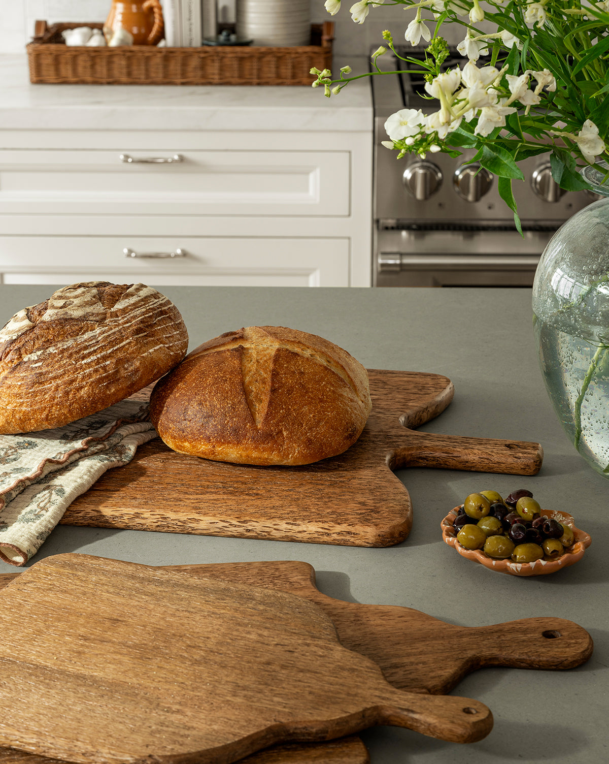 Two round loaves of bread rest on Longfellow Breadboards on a kitchen counter next to a small dish of olives. White cabinets and a vase of flowers add to the cozy kitchen decor in the background, by Vendixs