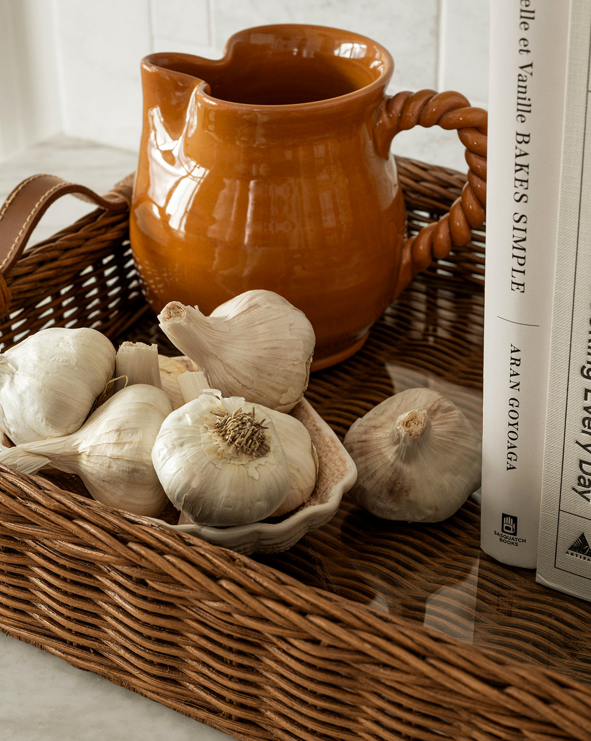 The Ashcroft Wicker Tray displays garlic bulbs in a shell-shaped dish, an orange ceramic pitcher with a twisted handle, and upright white cookbooks on a light glass countertop, by McGee & Co.