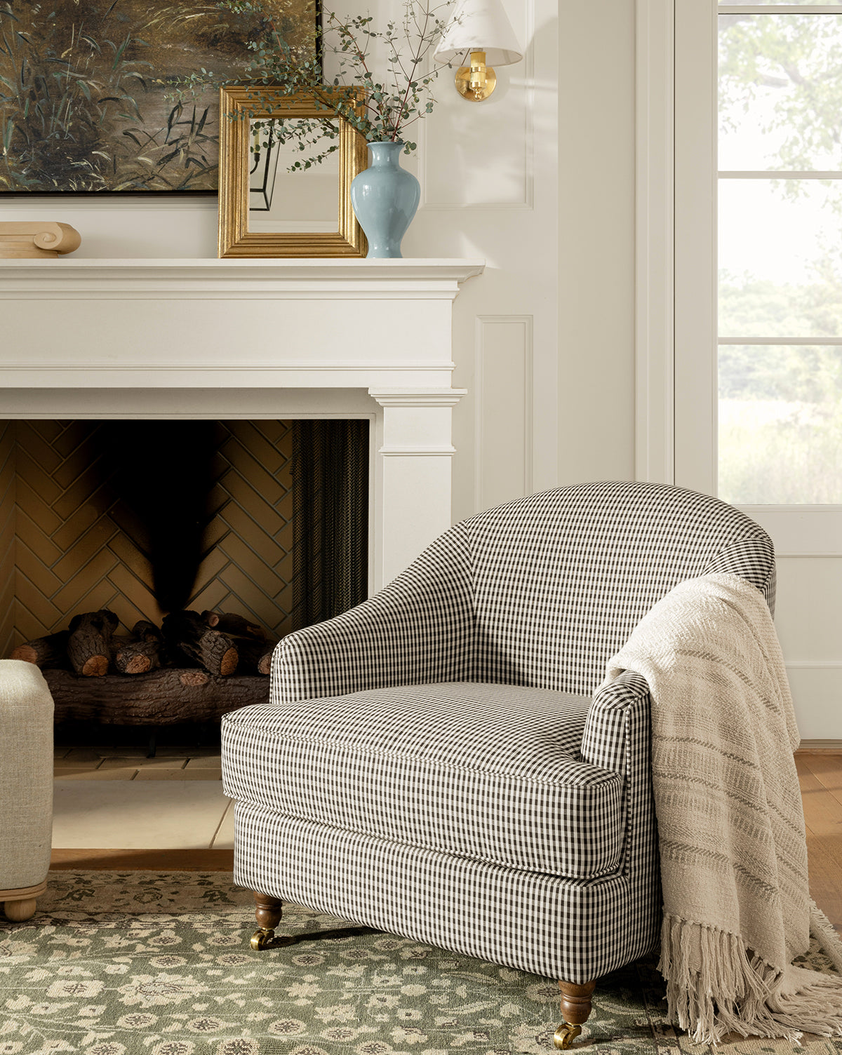 The Makerspalm Fletcher Lounge Chair, featuring a black-and-white checkered pattern, sits by the fireplace with a textured cream throw draped over one arm. Above the mantel are a vase, small frame, and nature painting by Vendixs