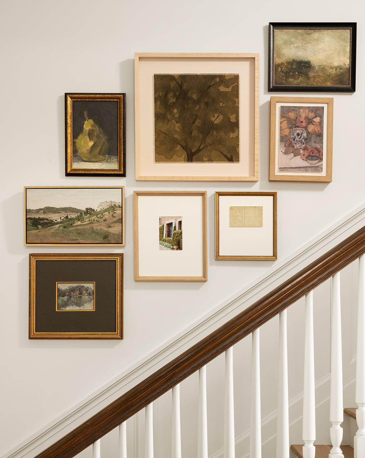 A gallery wall above a staircase displays seven framed art pieces, including the Blowing Brush print, arranged on a light wall above a white railing for an elegant and artistic accent, by Vendixs