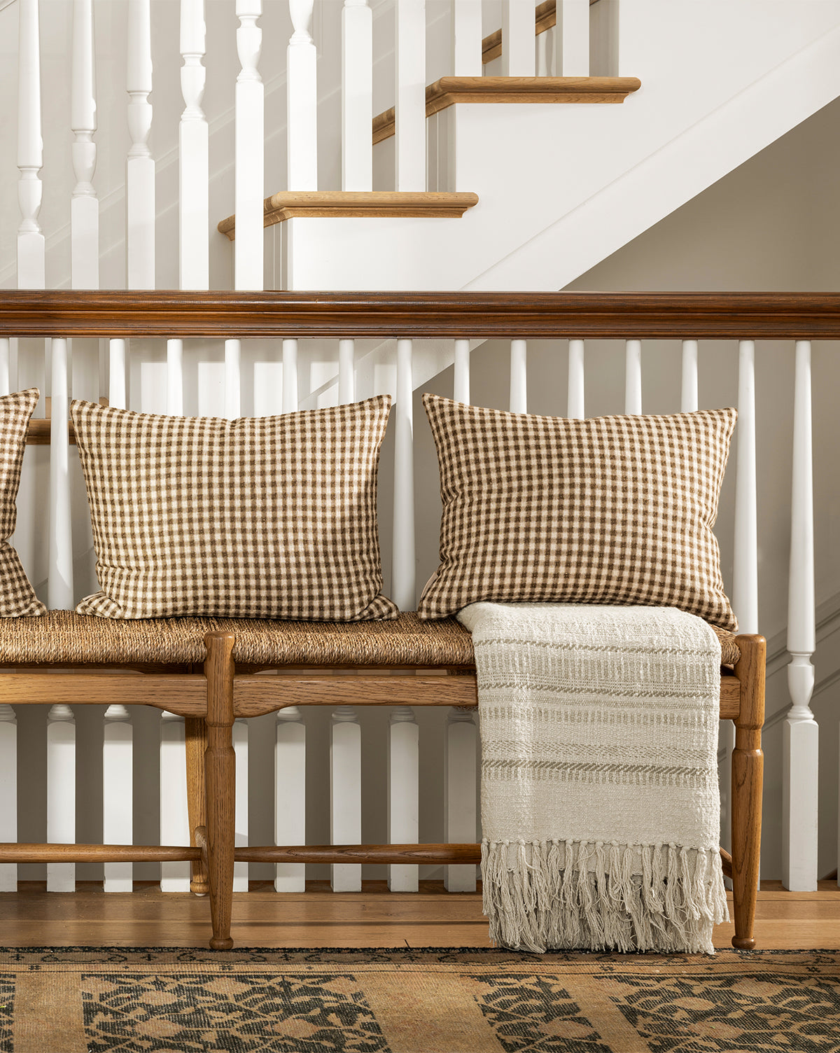A wooden bench with two brown gingham pillows and the Donahue Striped Throw by ILA HOME FASHIONS sits against a staircase with white spindles and wood accents, with a patterned rug on the floor below, styled by Studio Vendixs