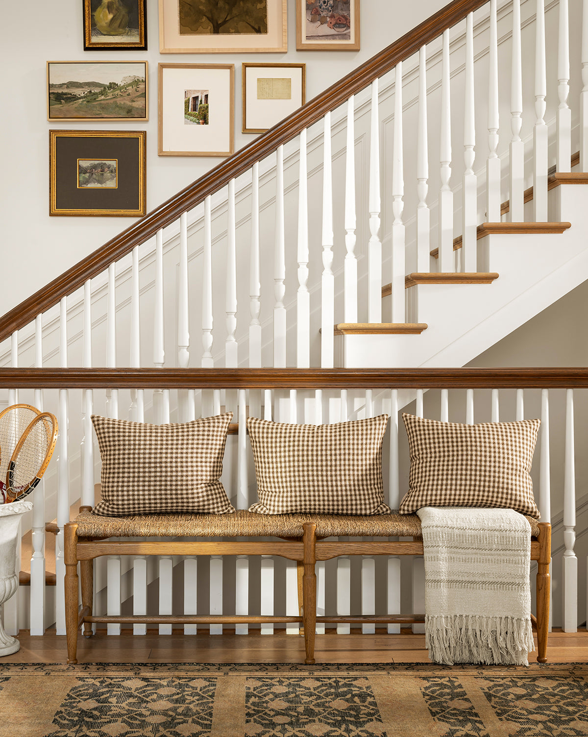 A cozy hallway features the Phipps Woven Bench topped with three plaid pillows and a fringed throw, set by a white staircase with art. A patterned rug and small table display vintage tennis rackets, while honey oak tones warm the space, by Vendixs