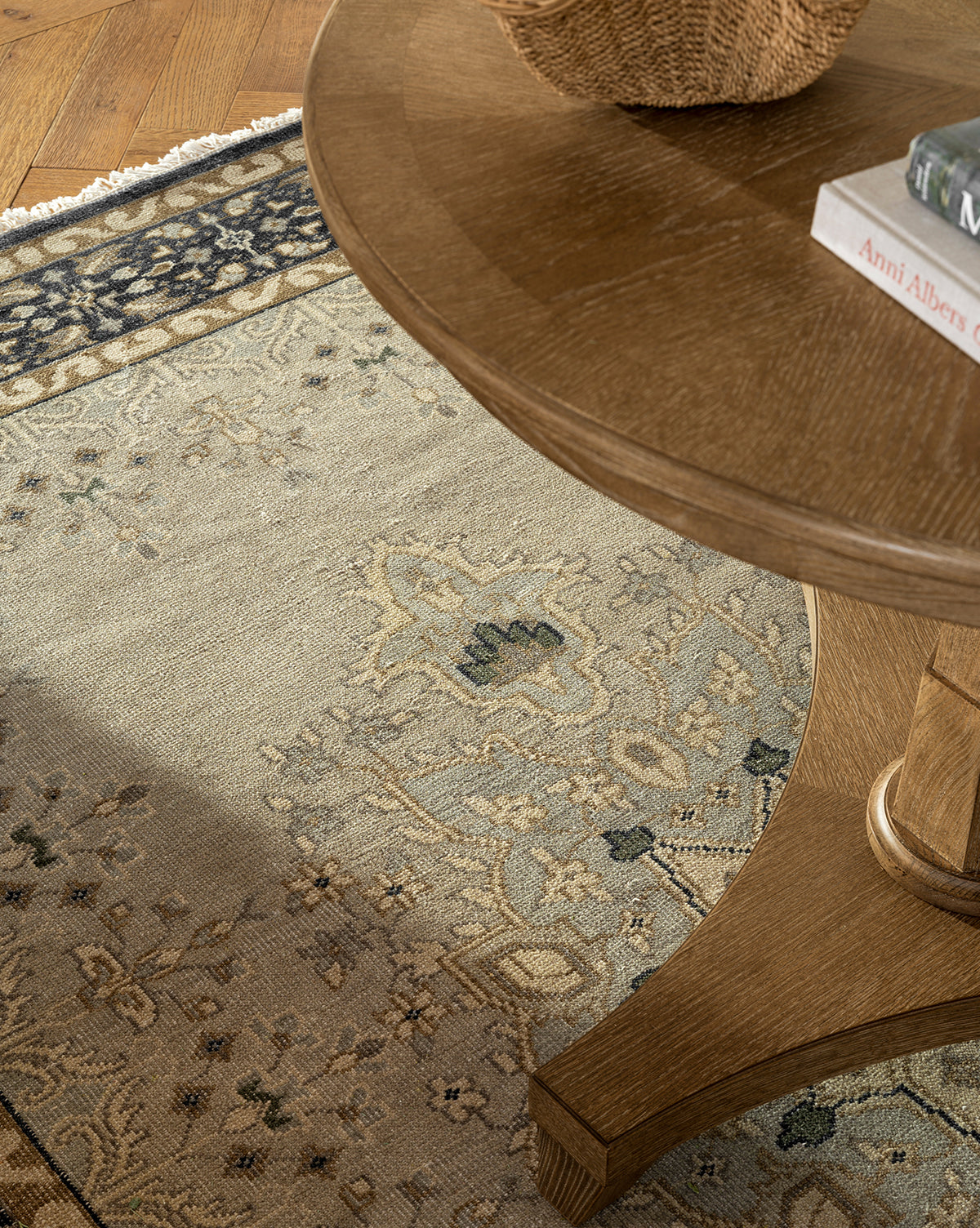 A close-up of a round wooden table on the Deveraux Hand-Knotted Wool Rug in beige and blue, with a woven basket and books on top. The table’s leg and the rug’s intricate design are clearly visible, by Vendixs