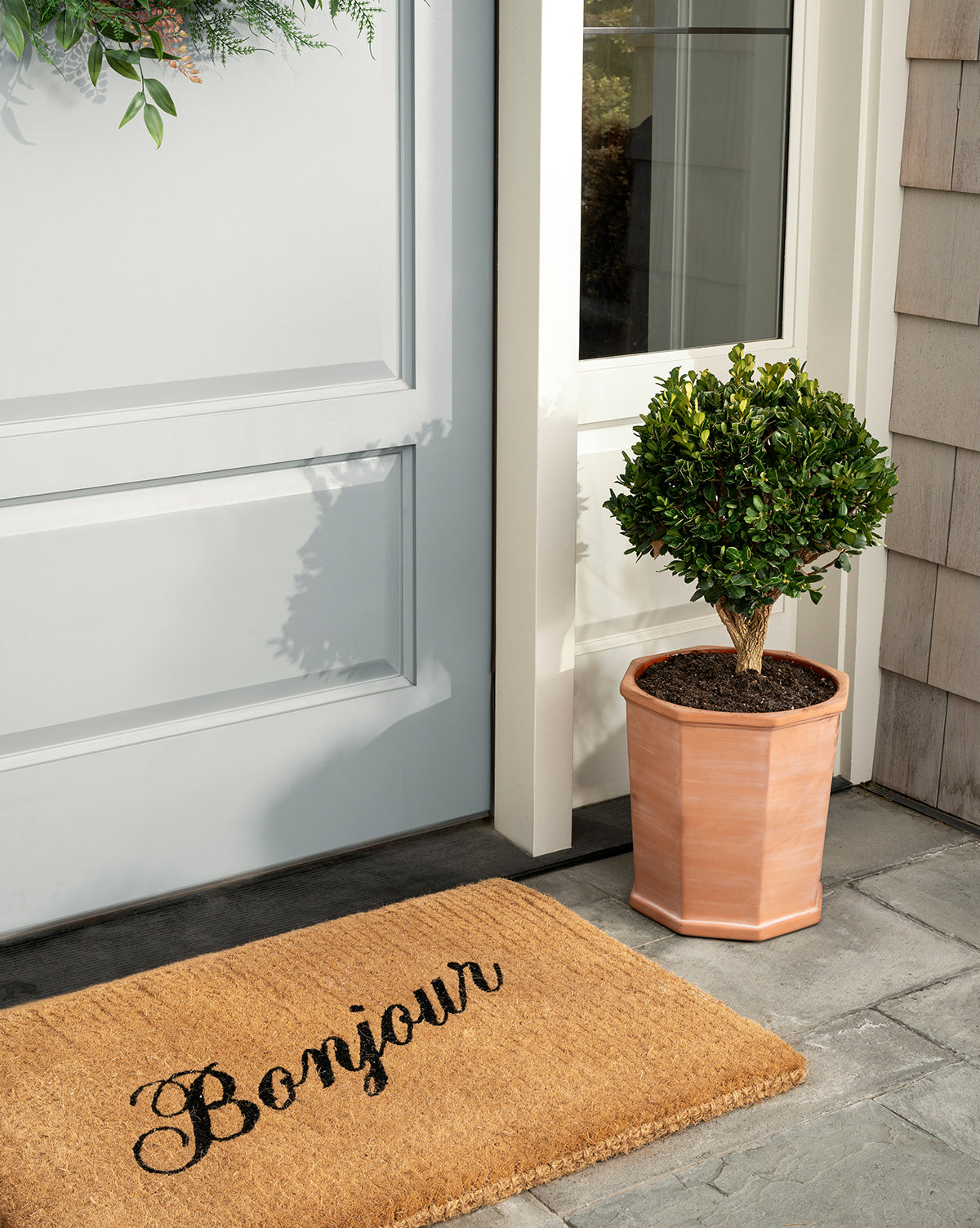 A handwoven coir Bonjour Doormat rests before a light blue door, bringing charm to your entryway, while a potted topiary in a light brown planter completes the welcoming scene, by McGee & Co.