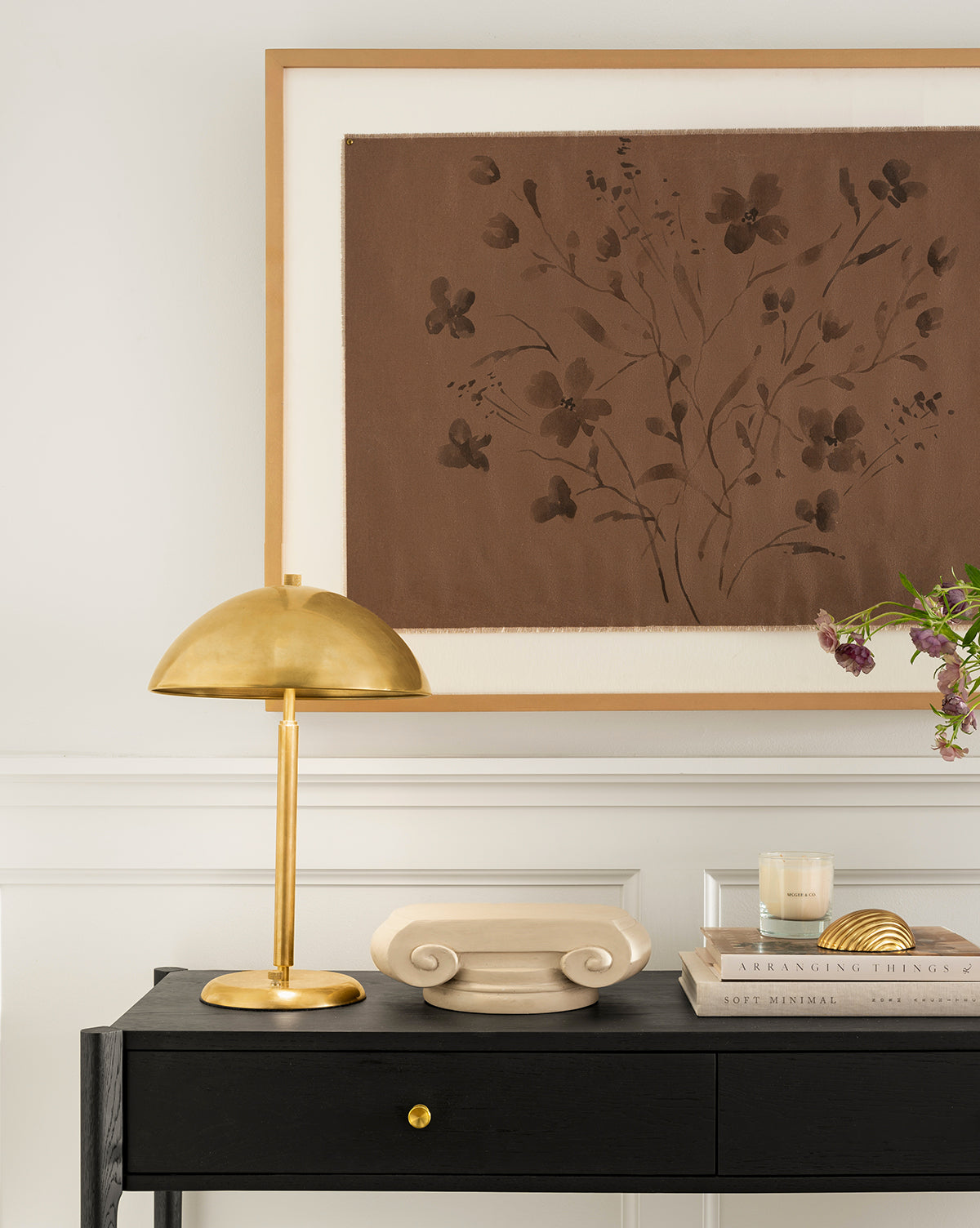 The Aldorno Desk Table Lamp sits on a black console table alongside a decorative scroll, books, candle, and glass vase with flowers, beneath brown floral artwork. This display enhances the living rooms elegance against a white wall, by Vendixs