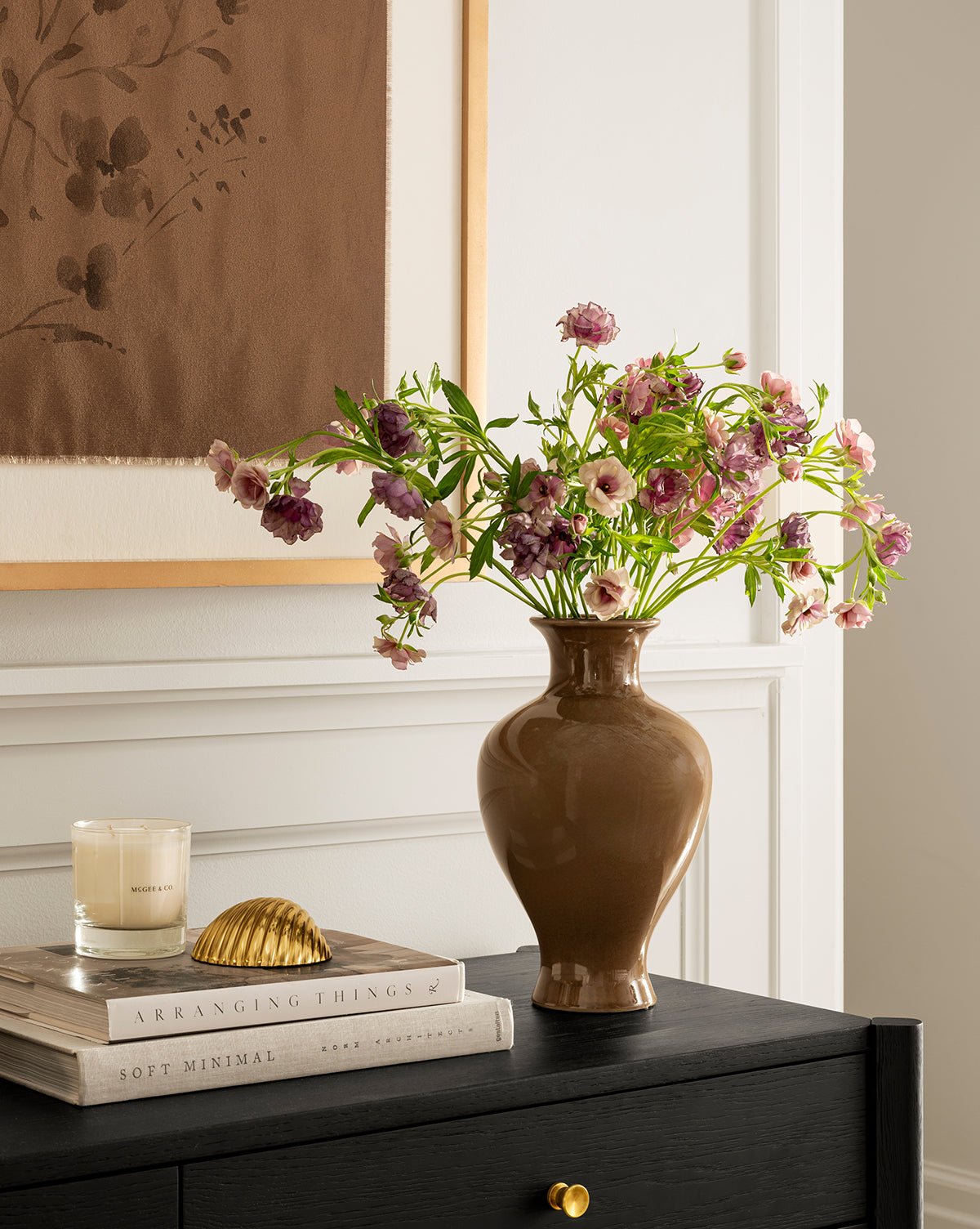 A brown Natalia Vase filled with pink and purple flowers sits on a black table next to a white candle, stacked books, and gold seashell decor, with framed artwork hanging on the wall in the background, by Vendixs