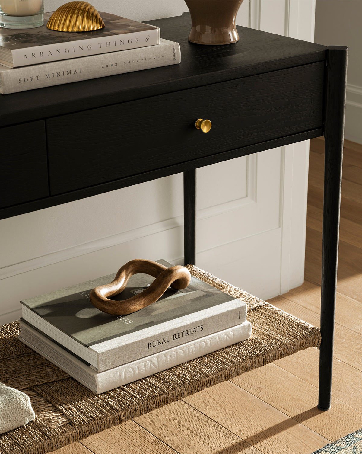 The Dana 2-Drawer Console in black with brass knobs stands on a wooden floor. Books and decor sit on top and a woven lower shelf, making it ideal for stylish entryways. Featured: Soft Minimal and Rural Retreats, by Vendixs