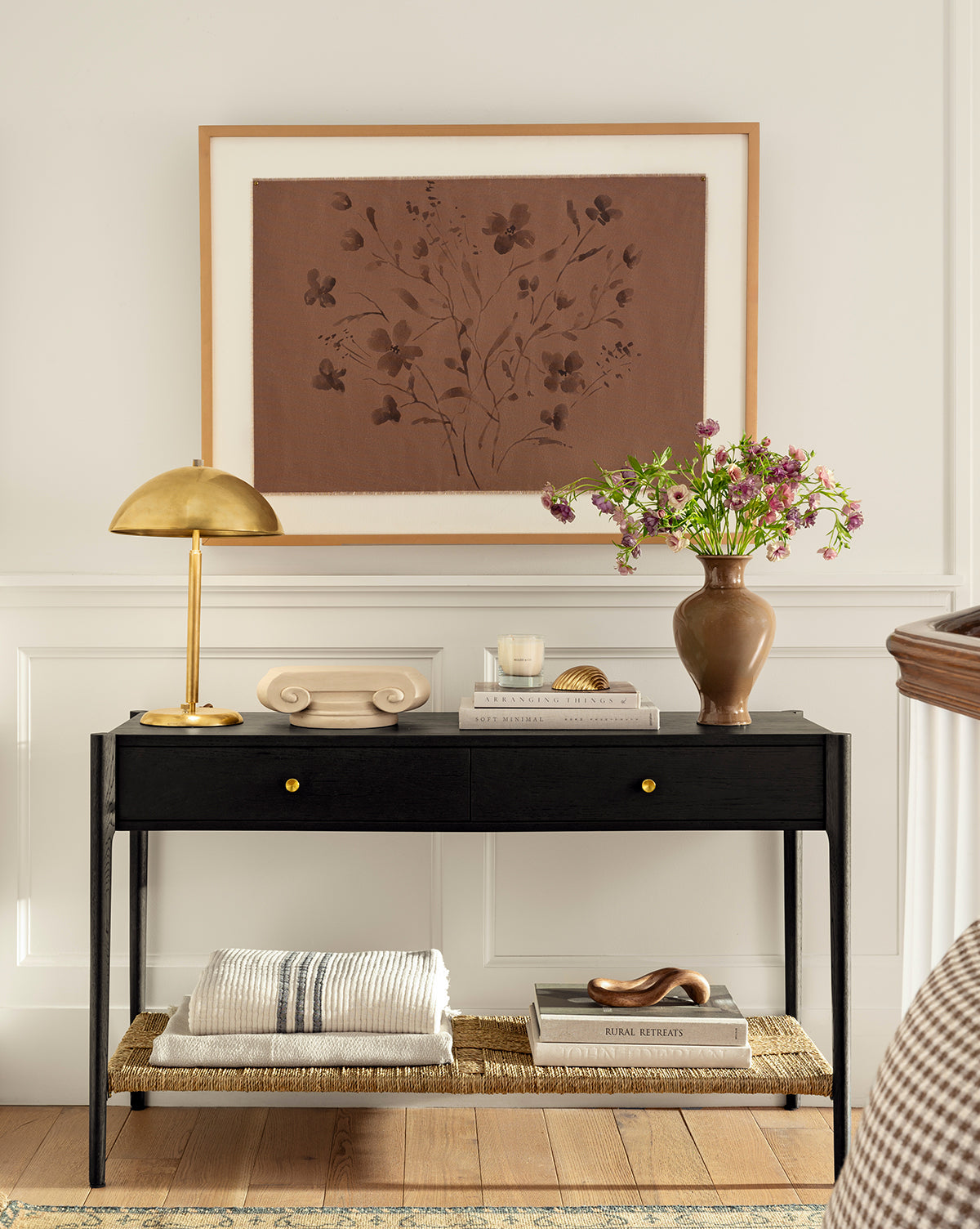 A Creative Co-op Anneliese Striped Throw is draped on a black console table with gold knobs, accented by books, a gold lamp, decor items, and a brown vase with pink flowers. Above hangs floral artwork in brown tones on a white paneled wall—ideal for luxury home decor. Vendixs