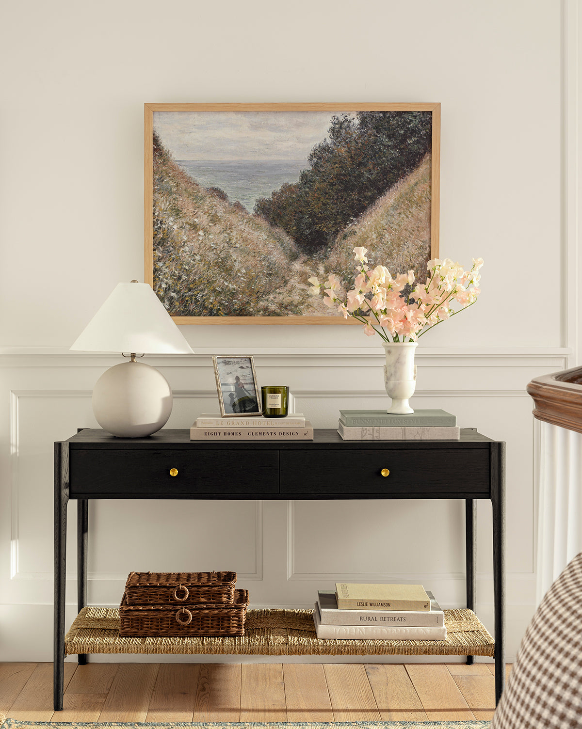 A black console table with two drawers displays a round white lamp, photo frame, books, and the Rayla Marble Vase filled with pink flowers; a landscape painting hangs above, with baskets and books on the lower shelf by Vendixs