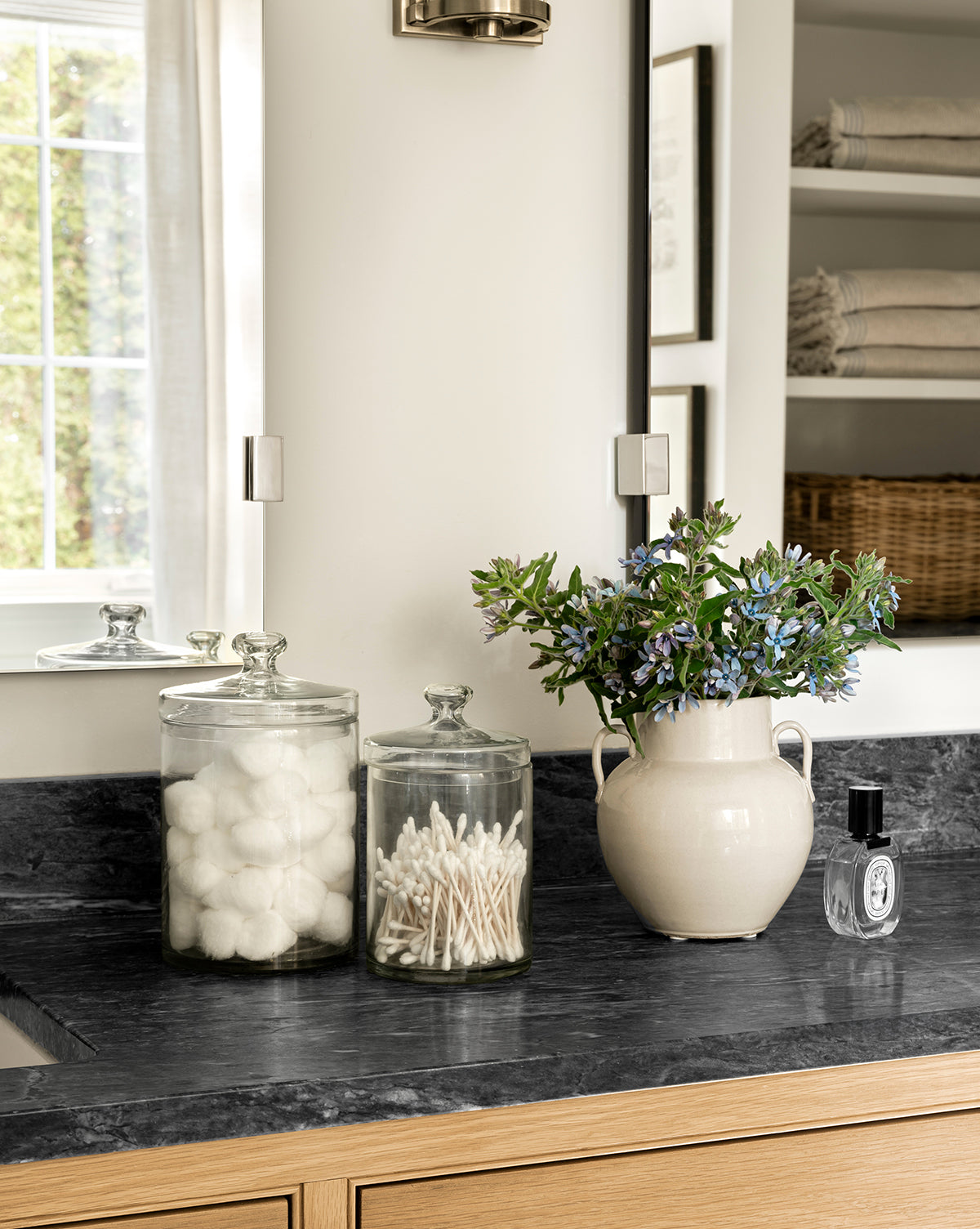 A Rasley Handled Jar sits on the bathroom counter beside glass jars of cotton balls and swabs, a cream vase with blue flowers, and a porcelain jar. Towels and a wicker basket are neatly stacked on shelves in the background, by Vendixs