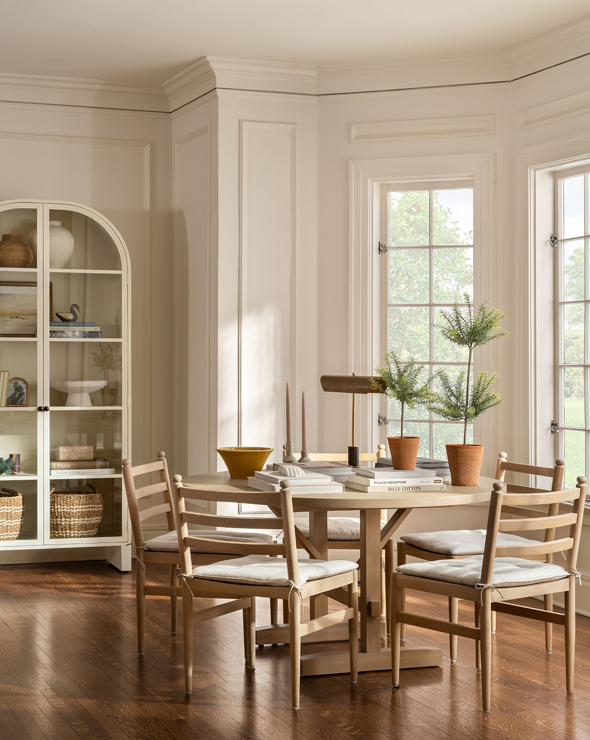 A bright, elegant dining room features a round white oak table, four Romany Dining Chairs with linen cushions, potted plants, books, and decorative objects. Large windows fill the space with light; a cabinet with shelves stands in the corner, by McGee & Co.