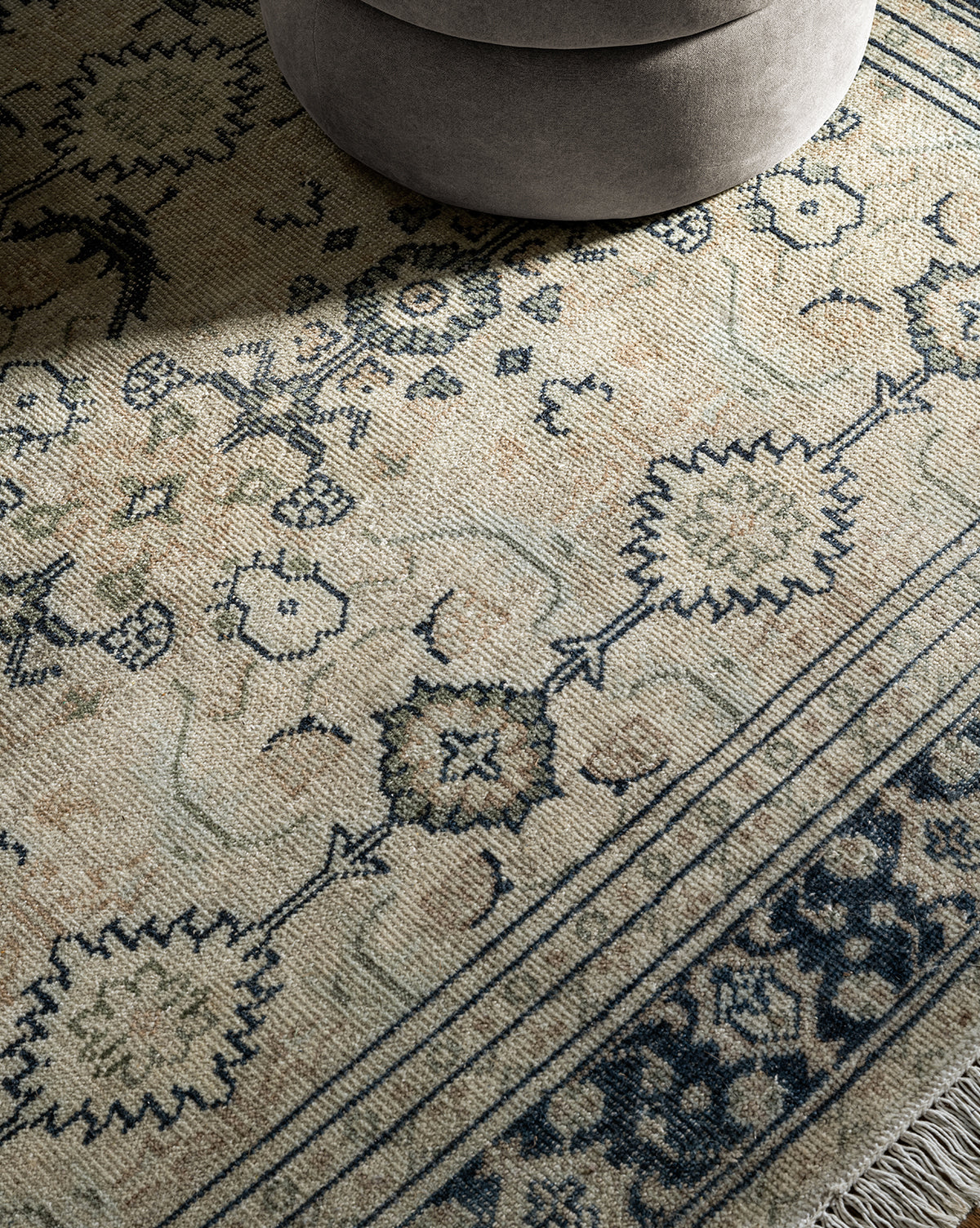 A close-up of the Vercelli Hand-Knotted Wool Rug, showing its intricate blue and beige patterns with tasseled edges, alongside part of a round light gray upholstered ottoman in the upper corner, by Vendixs