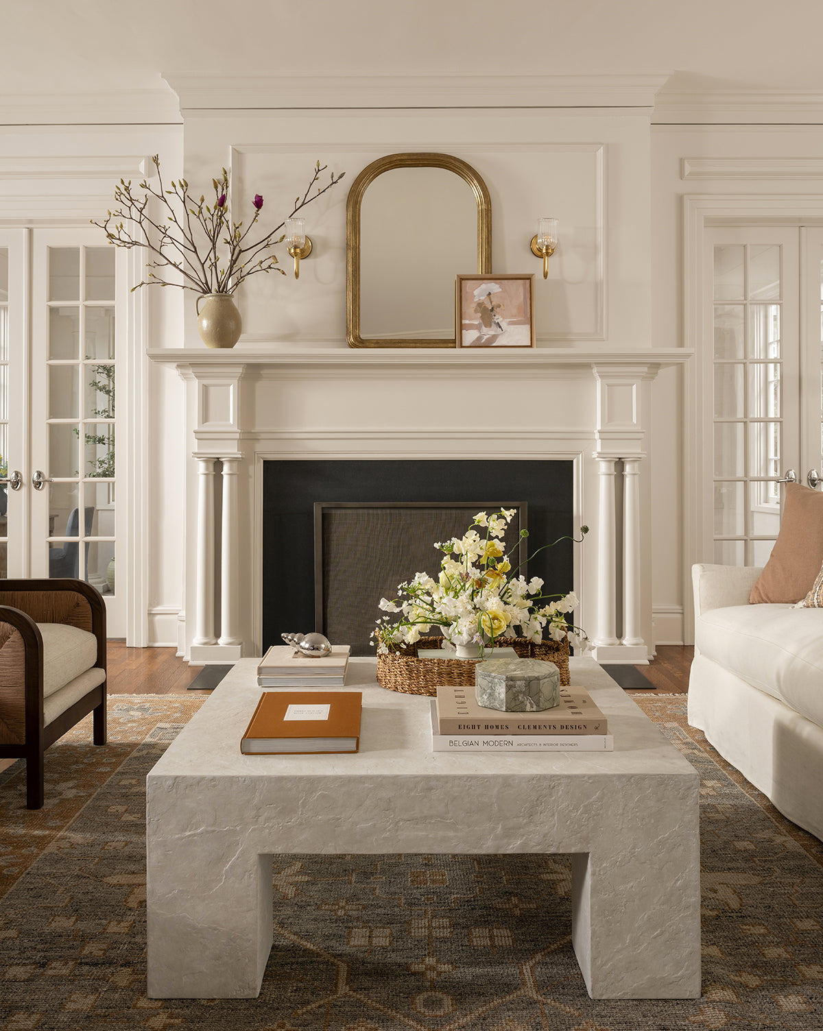 A bright, elegant living room with a stone coffee table, floral arrangements, books, a white sofa and armchair, and a white fireplace featuring the Giada Wall Mirror in its moulded frame with decorative objects above the mantel, by Vendixs