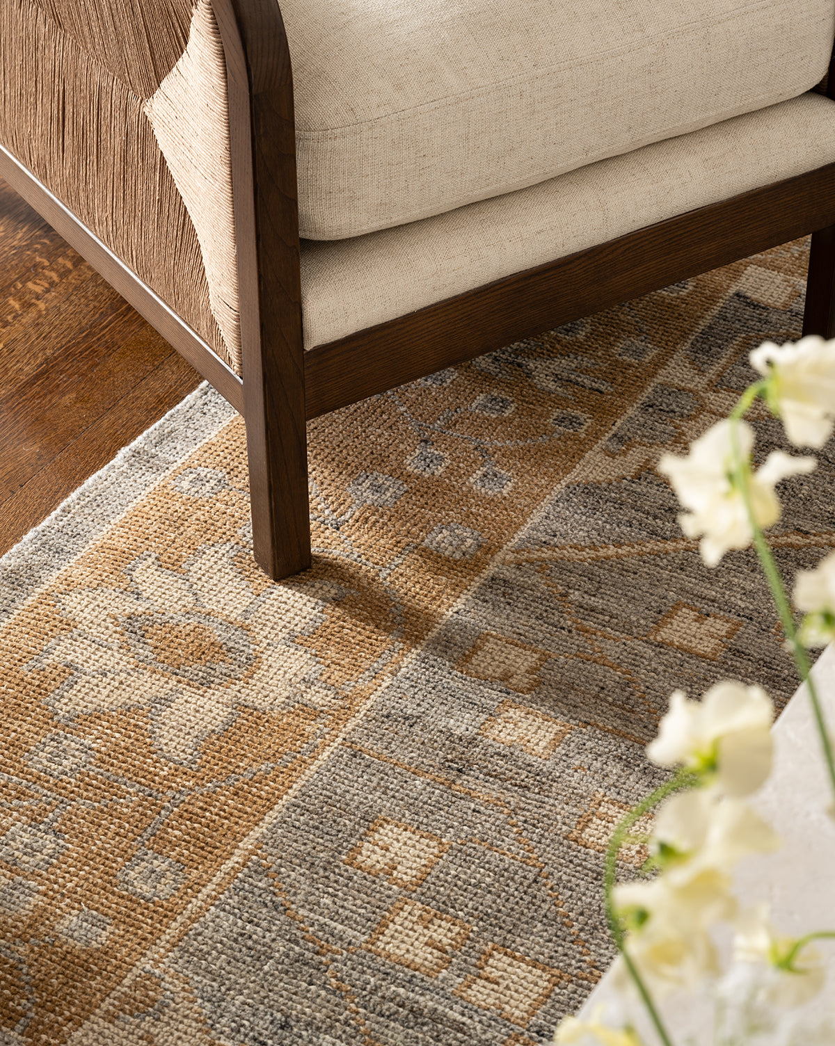 A close-up of a beige cushioned wooden chair on the Elison Hand-Knotted Wool Rug featuring gray, tan, and cream tones; white flowers are partially visible in the foreground, by Vendixs