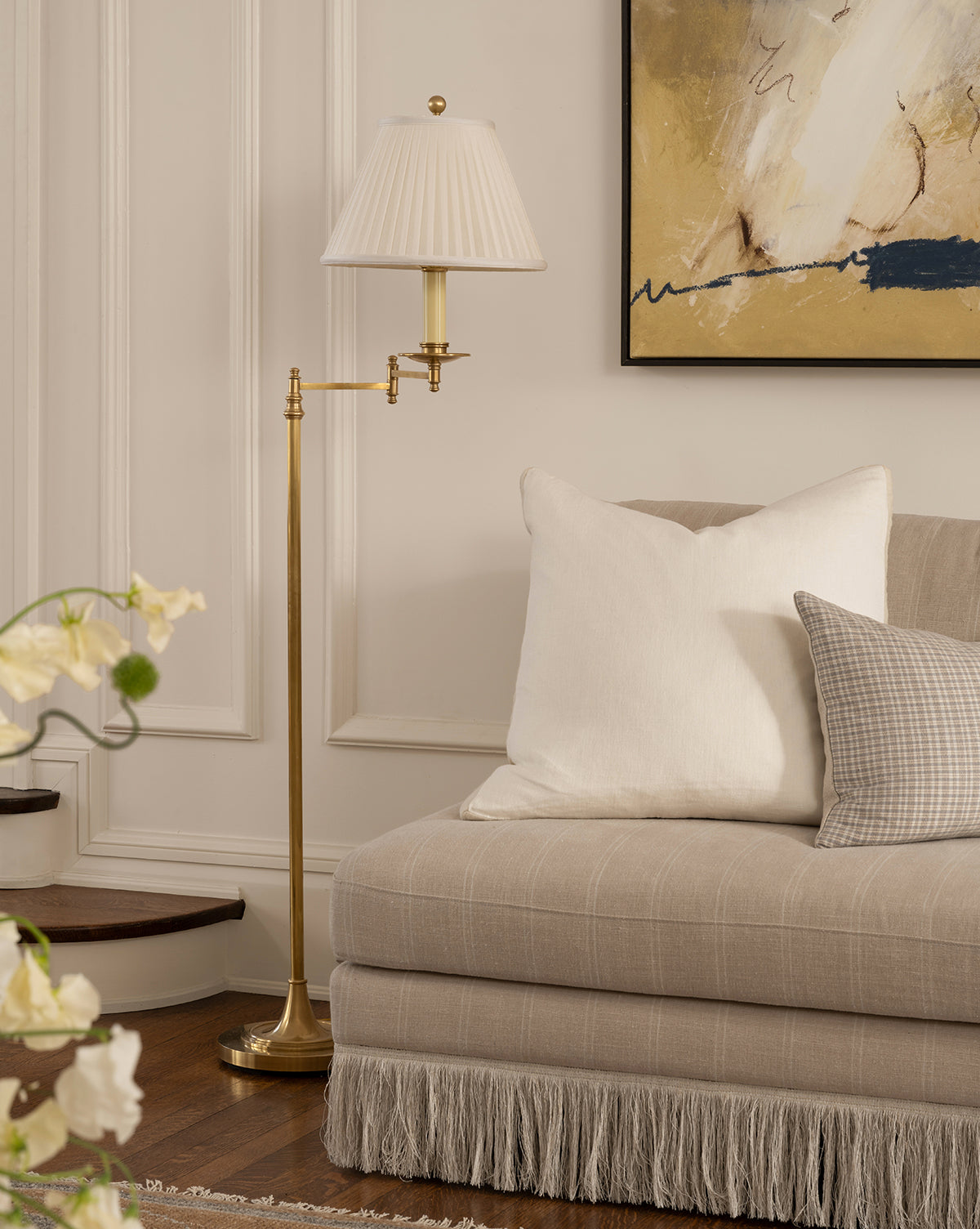 A cozy living room corner with a beige sofa, white and plaid pillows, abstract wall art, wooden stairs in view, and the Dorchester Swing Arm Floor Lamp featuring an antique brass finish and pleated silk shade, by Vendixs