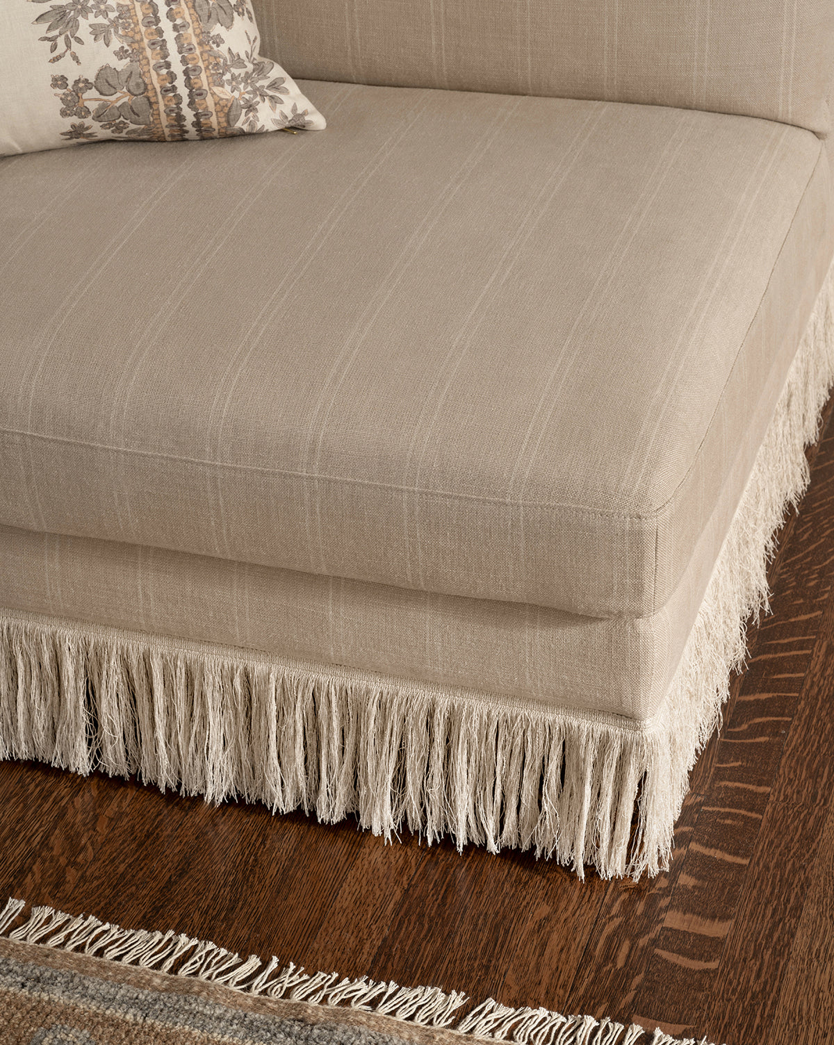 The Philippa Settee (Ready to Ship) in beige features decorative fringe along the bottom edge, set on a dark wooden floor. A patterned pillow rests on the settee and part of a woven rug is also visible, by Vendixs
