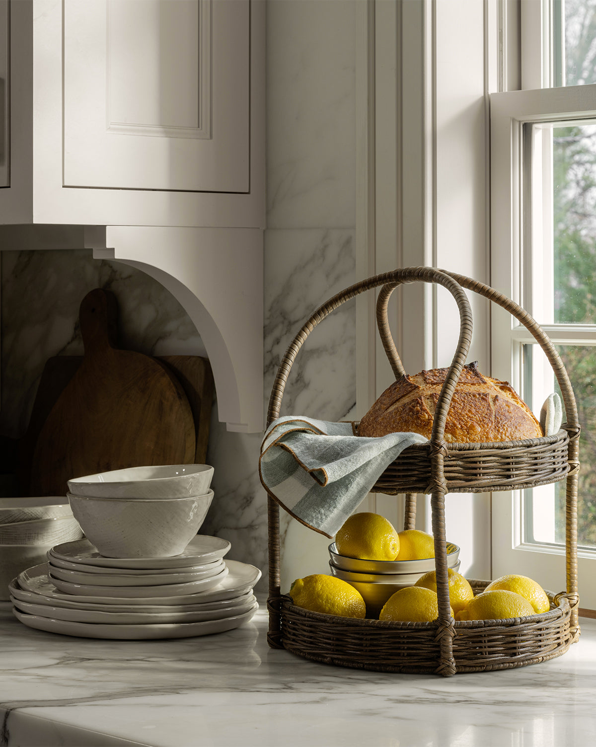 The Aneesa Two-Tiered Wicker Tray, filled with a loaf of bread, a folded cloth, and lemons, rests on a marble kitchen counter near stacked white dishes by a sunlit window by Vendixs