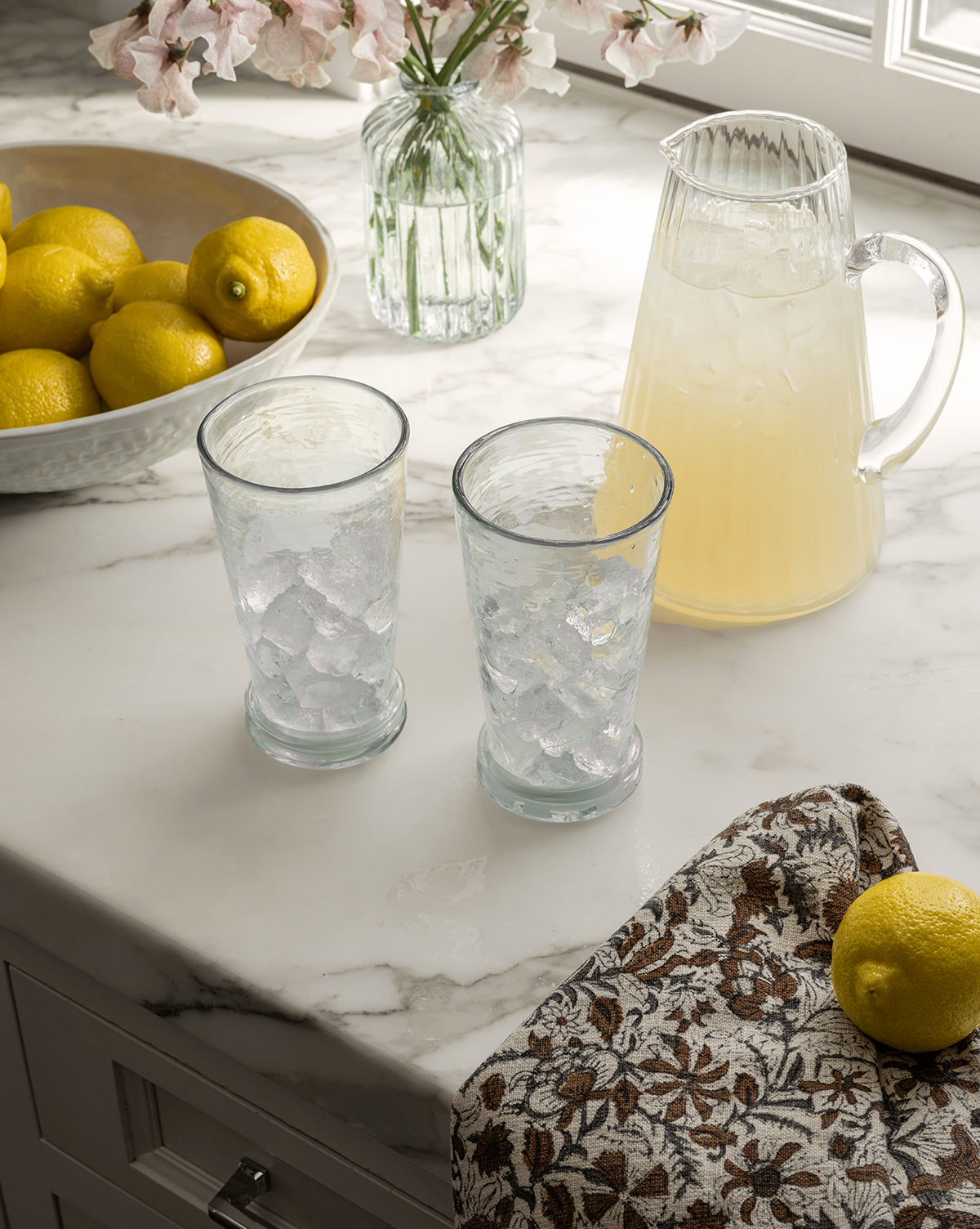 The Adonis Ribbed Glass Pitcher, filled with lemonade and paired with ice-filled glasses, fresh lemons, flowers, and a patterned cloth on a sunlit marble countertop, creates an effortlessly stylish kitchen scene, by Vendixs