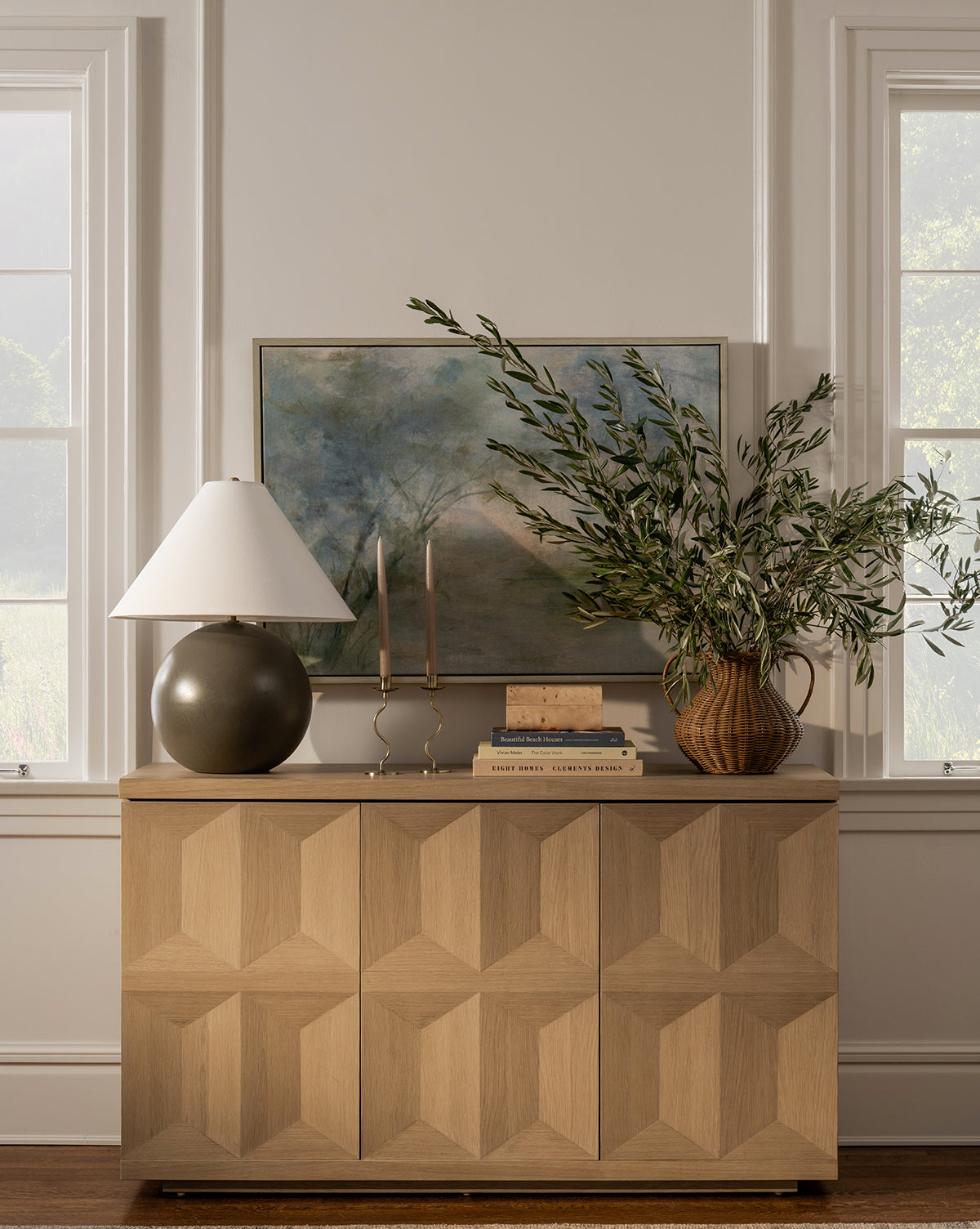 A light wood cabinet with a geometric pattern stands between two windows. On top are the Tess Large Ceramic Table Lamp with a white shade, two candlesticks, stacked books, and a vase of greenery. A landscape painting hangs on the wall behind by Vendixs
