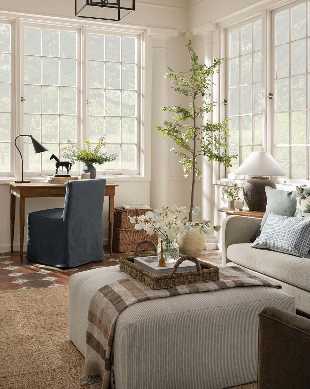 A bright, airy living room with large windows, a beige sofa, a Rowe Fine Furniture Culpepper Ottoman with tray, a small desk and blue chair, plus decorative plants and flowers throughout. Vendixs
