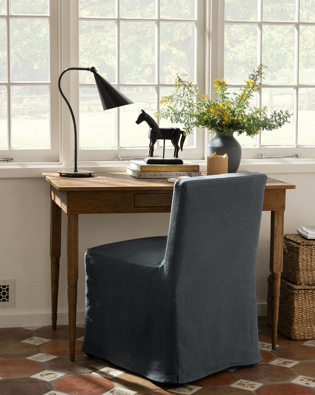 An Olivier Slipcover Dining Chair faces large windows at a wooden desk topped with a black lamp, stacked books, a black horse figurine, and a vase of yellow flowers. Wicker baskets sit nearby on the floor, by McGee & Co.