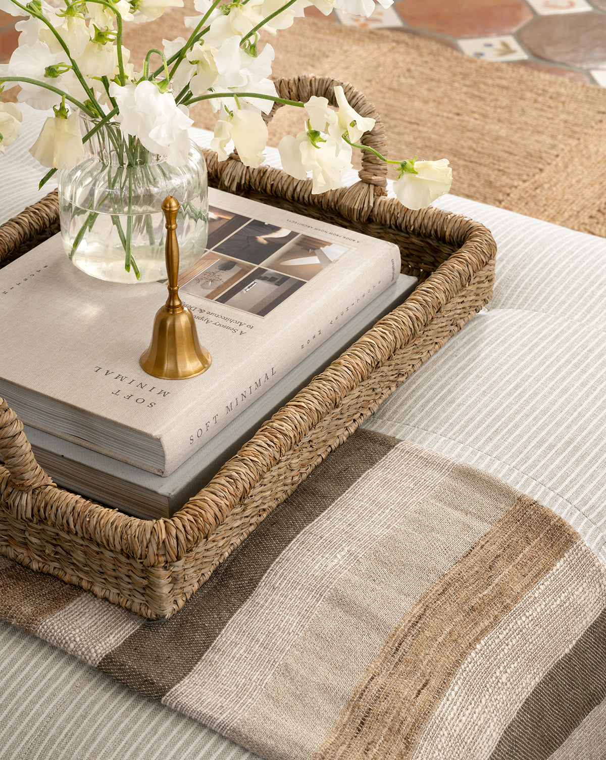 The Elsher Bell, paired with a book, brass candle snuffer, and glass vase of white flowers, rests on a striped ottoman in a cozy room with soft neutral tones, by Vendixs