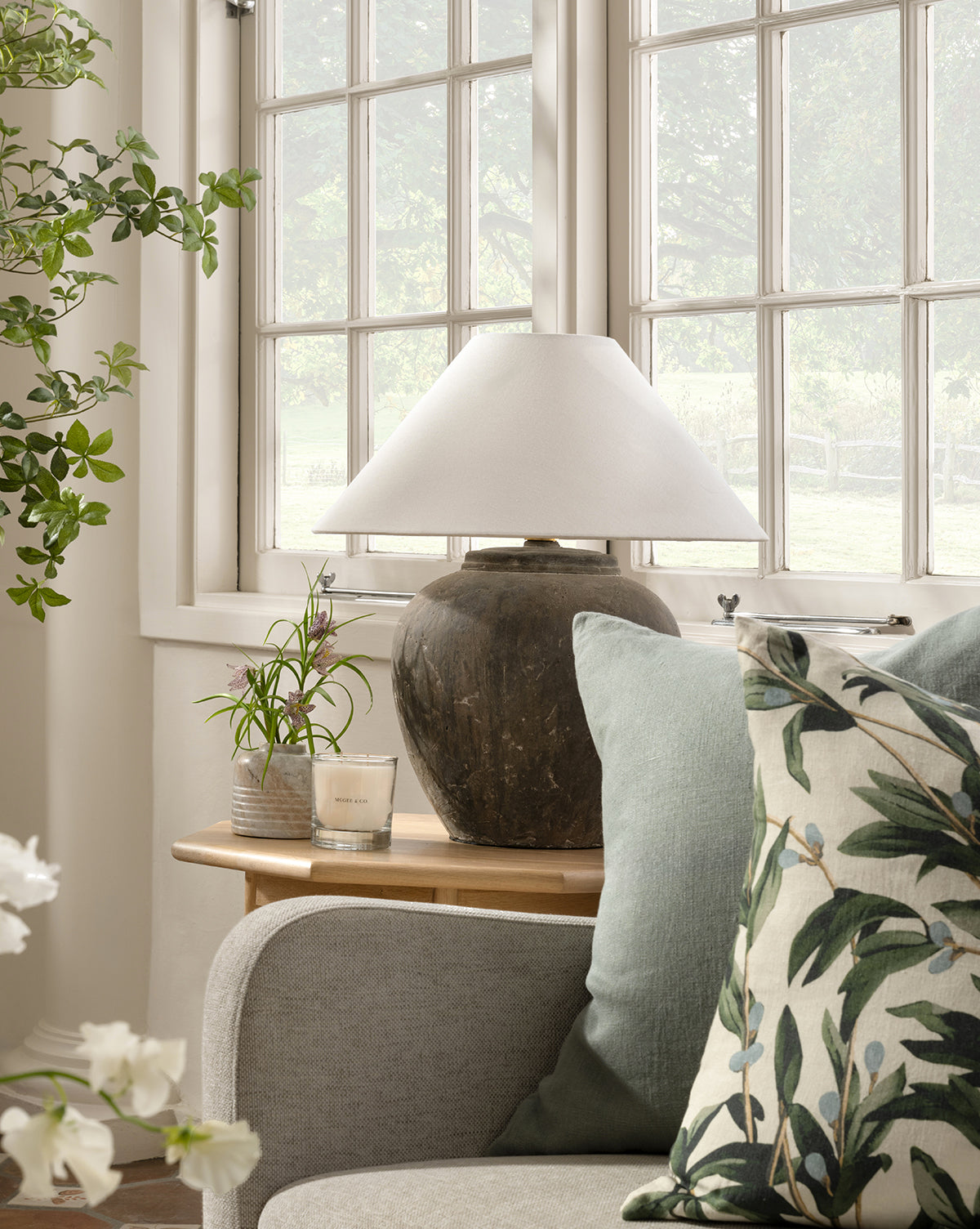 A cozy corner with a sofa, leafy pillow, light blue cushion, and wooden side table topped with a rustic lamp, Stinson Marble Bud Vase, small plant, and candle—set by large sunlit windows framing green outdoor foliage, by Vendixs