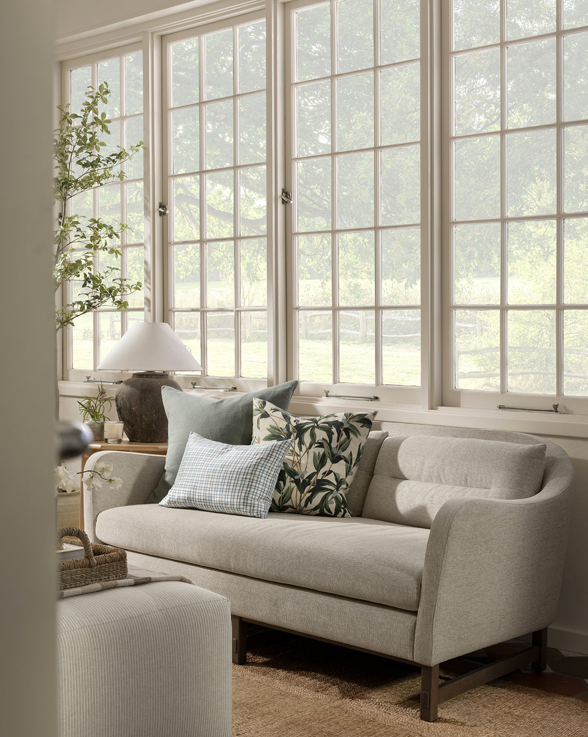 The Commodore Settee 68 in light gray twine crypton upholstery, with patterned and solid pillows, sits by large windows in a bright living room with greenery, a table lamp, and neutral decor as sunlight streams through the trees outside, by McGee & Co.