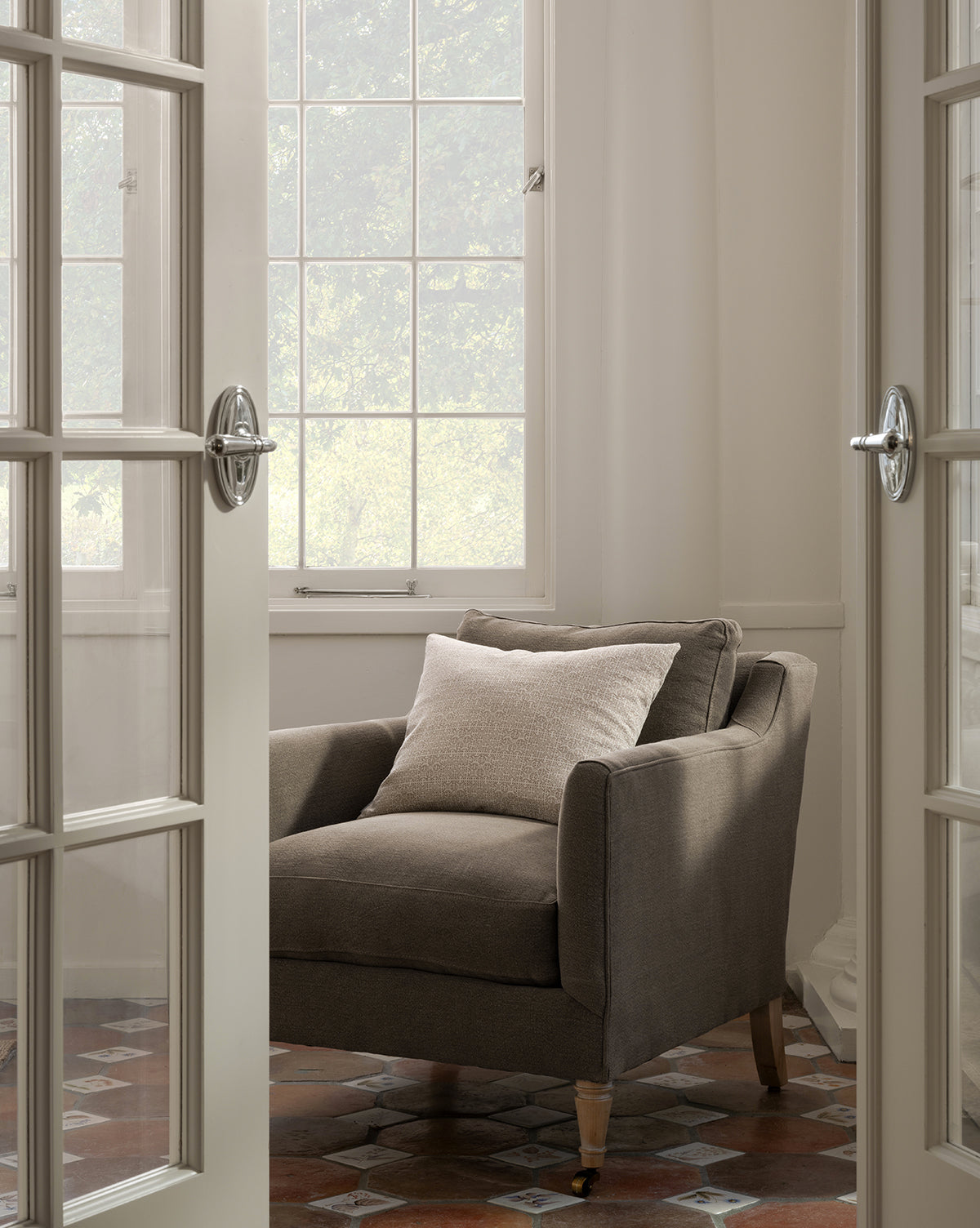 The Haverford Upholstered Lounge Chair with a light pillow sits in a sunlit corner near large windows, framed by open glass French doors on a patterned tile floor, by McGee & Co.