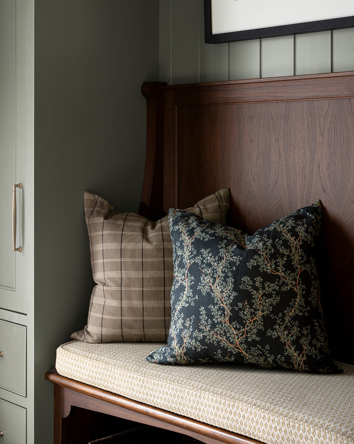 A wooden bench with a patterned seat cushion holds two pillows: one plaid and brown, the other dark with a botanical motif like the Antigone Pillow Cover. Nearby, there’s a cabinet and part of a framed picture in view, by Vendixs