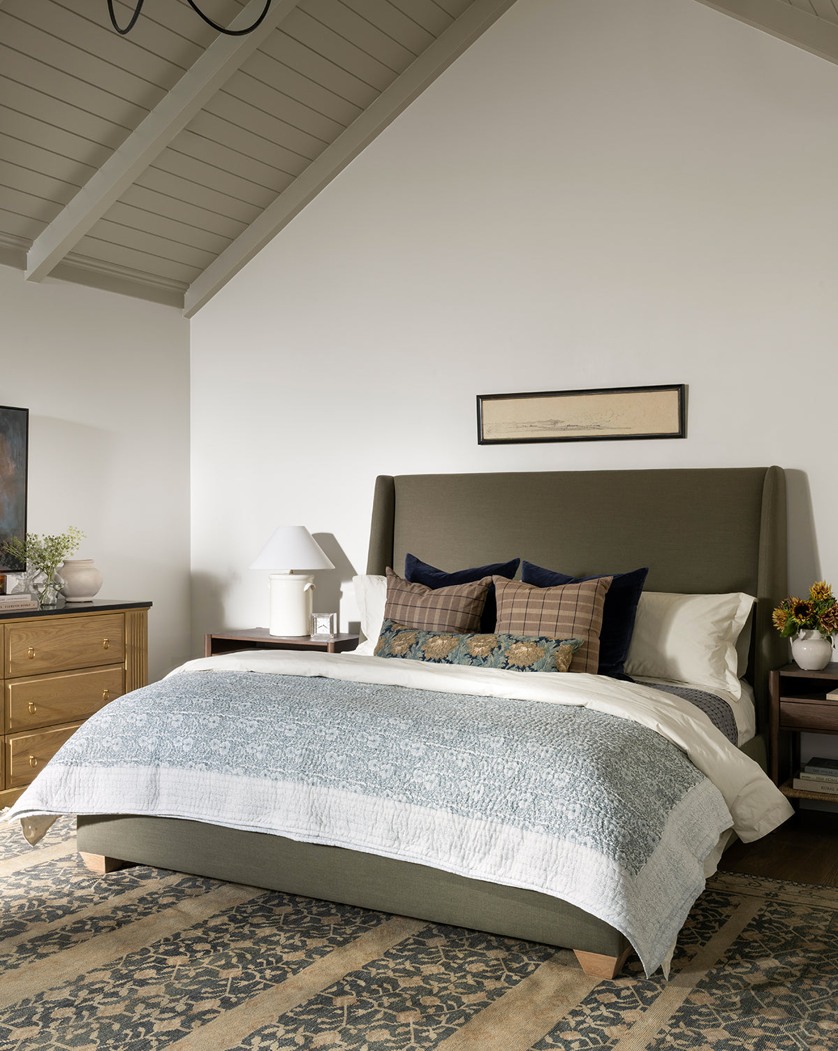 A cozy bedroom features a green upholstered bed with blue and plaid pillows and an artisanal bedding set, topped by the Blue Floral Block Print Quilt. Wooden nightstands hold lamps and flowers, with a dresser left under the vaulted gray ceiling, by Vendixs