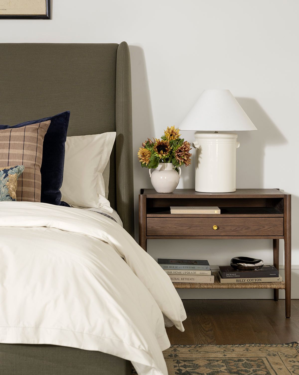 A cozy bedroom scene with a green upholstered bed, white bedding, and a Dana Walnut Oak Nightstand with brass hardware. On the nightstand are a white lamp, a sunflower vase, and stacked books on the lower shelf by Vendixs