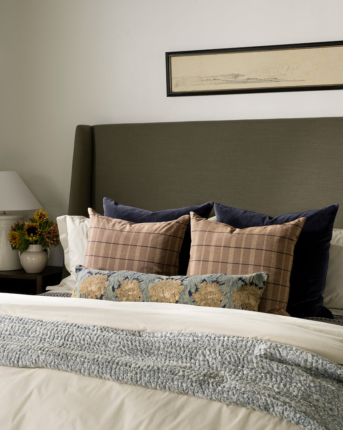 A neatly made bed with layered pillows, including a decorative pillow in the Bellevue Velvet Pillow Cover, a textured throw blanket, a potted plant and lamp on the bedside table, and rectangular framed artwork above the headboard, by Vendixs
