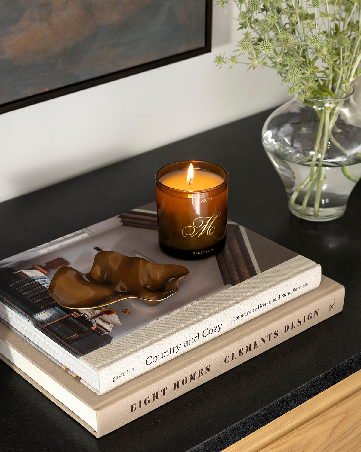 A lit brown candle rests on stacked books beside a Chiffonner Metal Object with a sleek metallic finish and a glass vase of green foliage, all arranged on a dark tabletop for a modern artistic display, by Vendixs