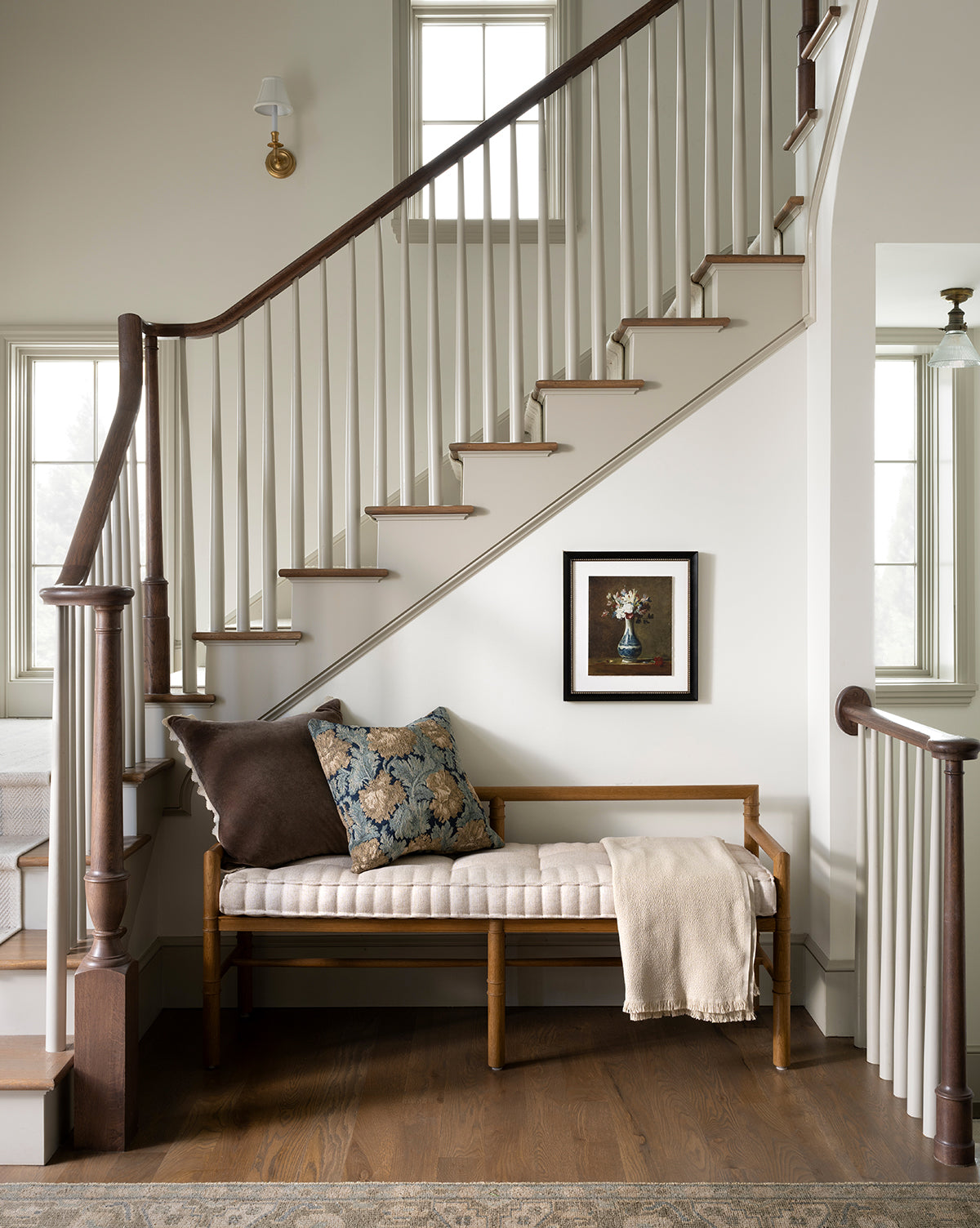 The Hindley Bench with upholstered cushions and a throw blanket sits beneath a staircase with white spindles and dark wood handrails. A framed flower print decorates the wall, as two windows brighten the space with natural light, by Vendixs