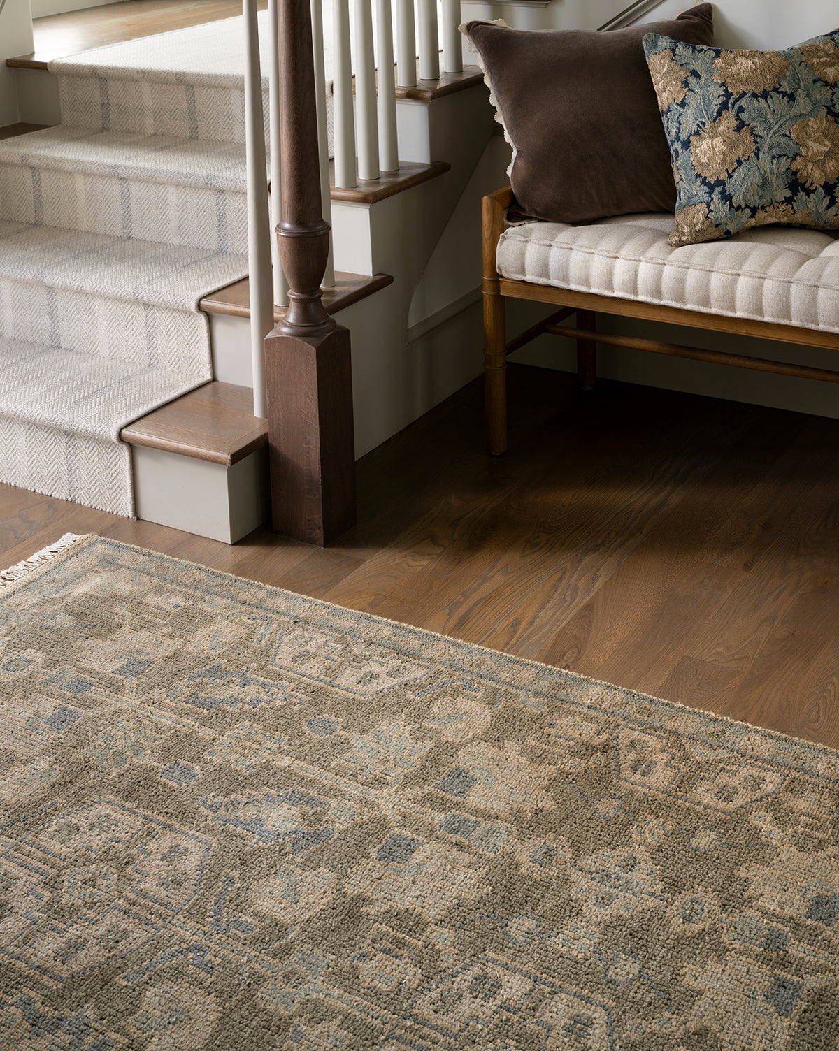 The Chevalier Hand-Knotted Wool Rug in neutral tones lies on the wooden floor near a staircase with carpeted steps. A wooden bench with a beige cushion and pillows beside the stairs adds timeless elegance to the space, by Vendixs