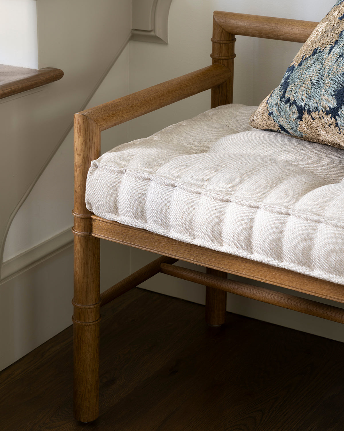 A close-up of the Hindley Bench features a light upholstered cushion and a decorative blue patterned pillow, set on a dark wood floor by a window and cream-colored wall, by Vendixs