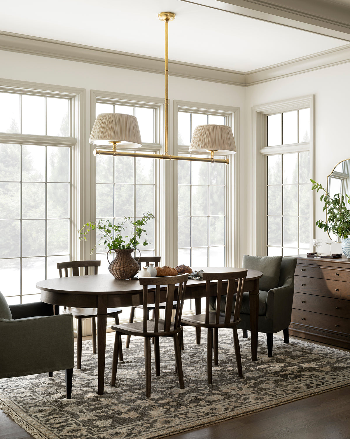 A bright dining room with large windows, a round dark wood table surrounded by Hemingway Chairs, a patterned rug, green armchairs, a vase of greenery, a sideboard, and a modern gold chandelier with two lampshades, by Vendixs