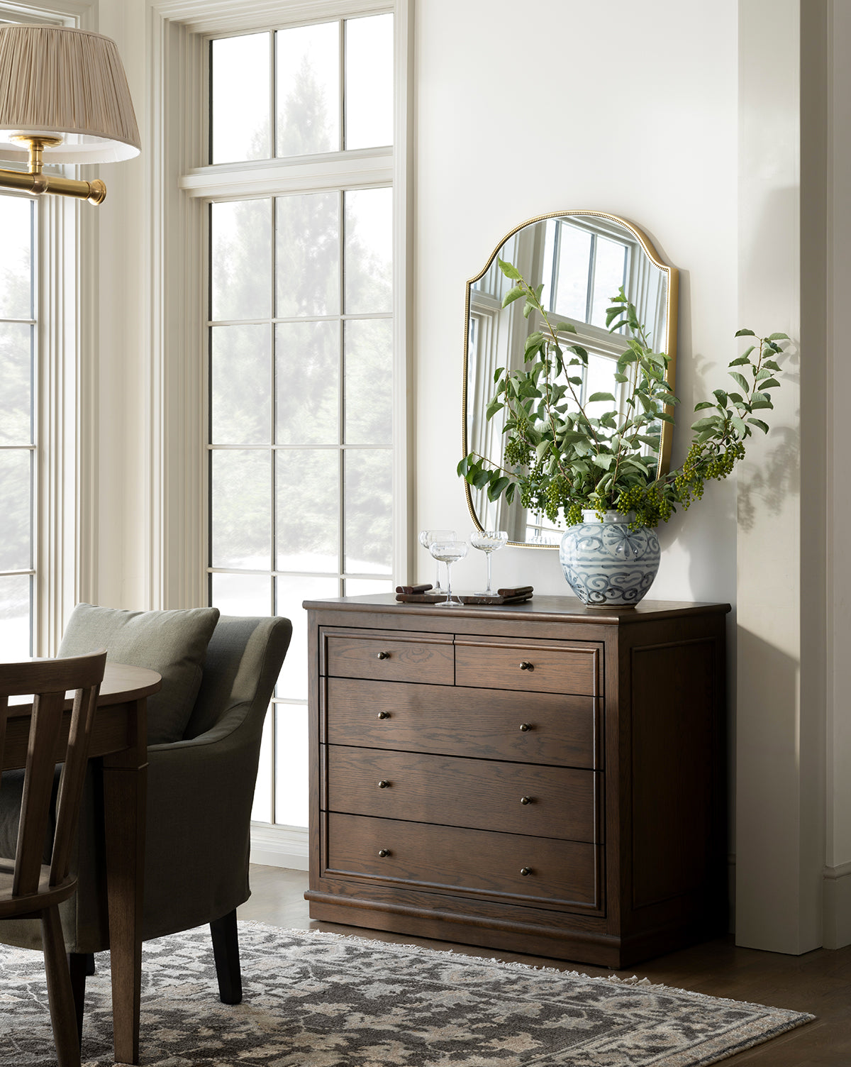 A wooden dresser with six drawers stands against a white wall beneath a gold-edged mirror. The Wilfred Vase with leafy branches sits on top next to two wine glasses, while large windows fill the space with natural light, by Vendixs