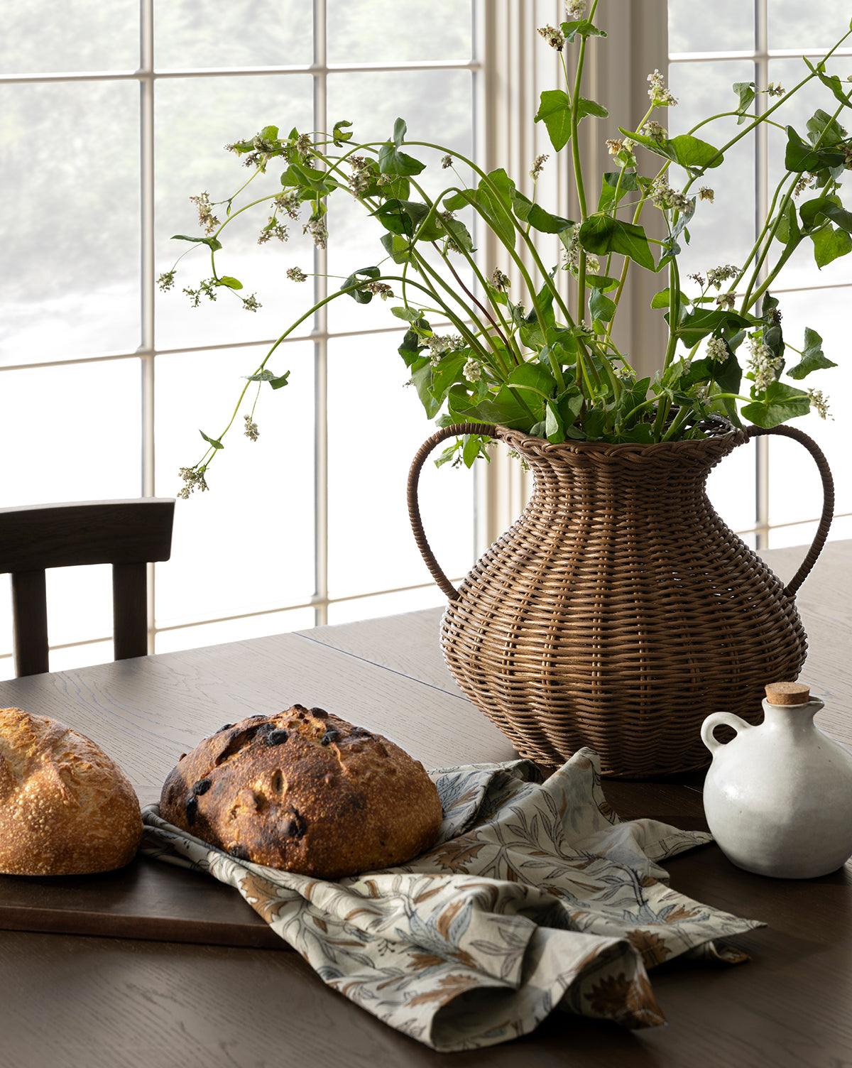 A wooden table is set with two loaves of bread on a cutting board, a patterned cloth, a white ceramic jug, and the Antonia Woven Vase filled with green leafy branches, in front of large sunlit windows, by Vendixs
