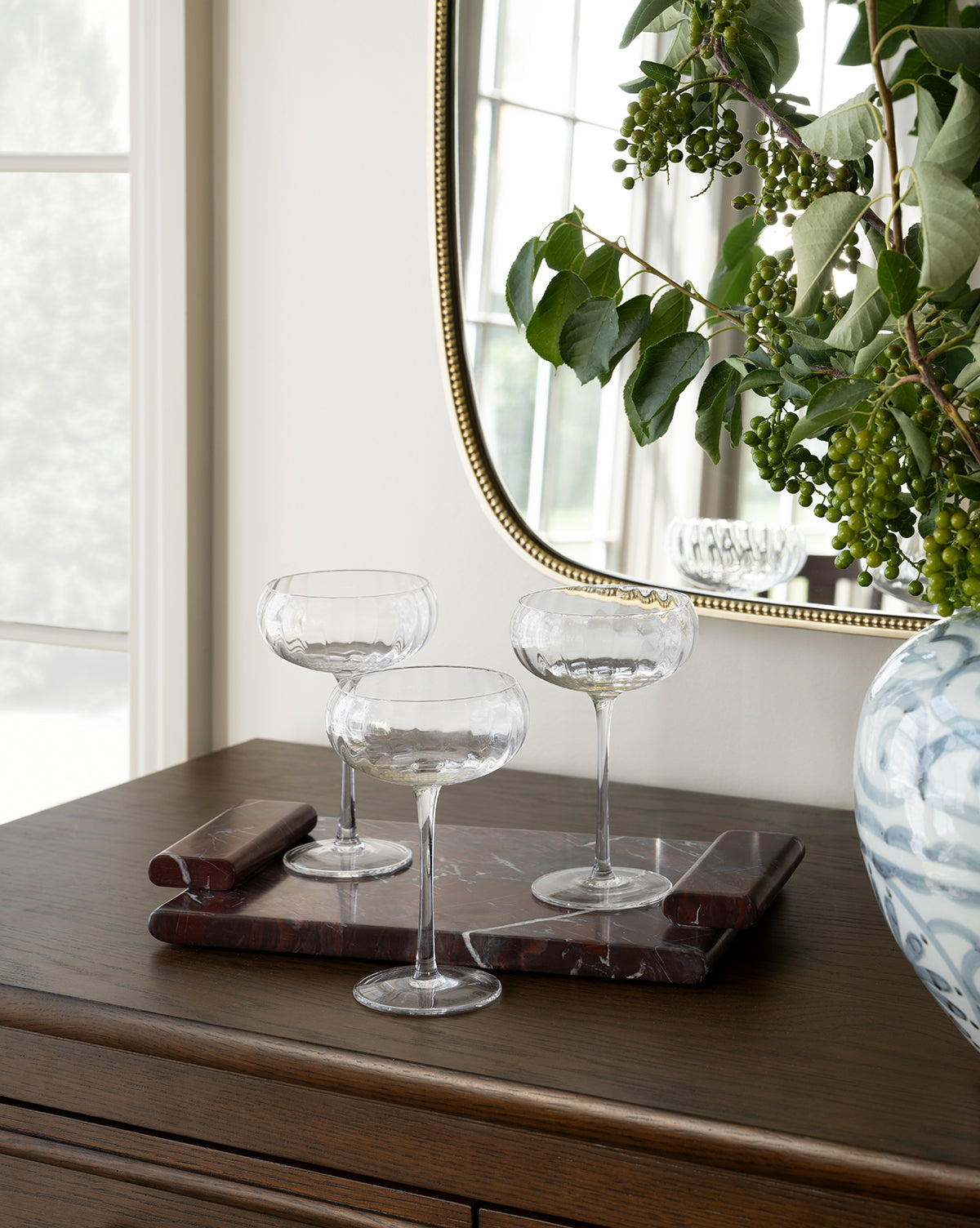 Three empty coupe glasses rest on the Auburn Marble Tray atop a wooden dresser, next to a large vase with green foliage. A mirror reflects the scene and a bright window glows in the background, by Vendixs