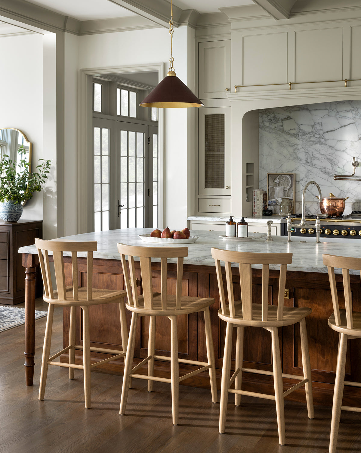 A modern kitchen with a marble island, Hemingway Stools, a copper pendant light, and neutral cabinets. The island features a bowl of apples and soap dispensers, while a potted plant rests near the counters in the background, by Vendixs