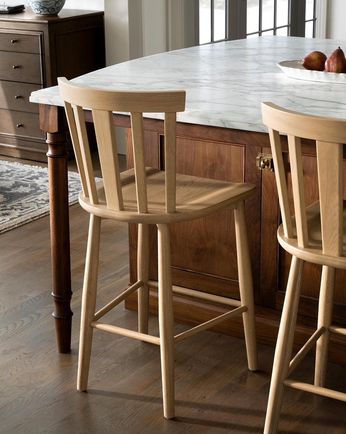A Hemingway Stool made from solid wood with a slatted backrest stands by a marble-topped kitchen island, complementing the wooden floor and cabinetry. A second Hemingway Stool is partially visible near the kitchen counters, by Vendixs
