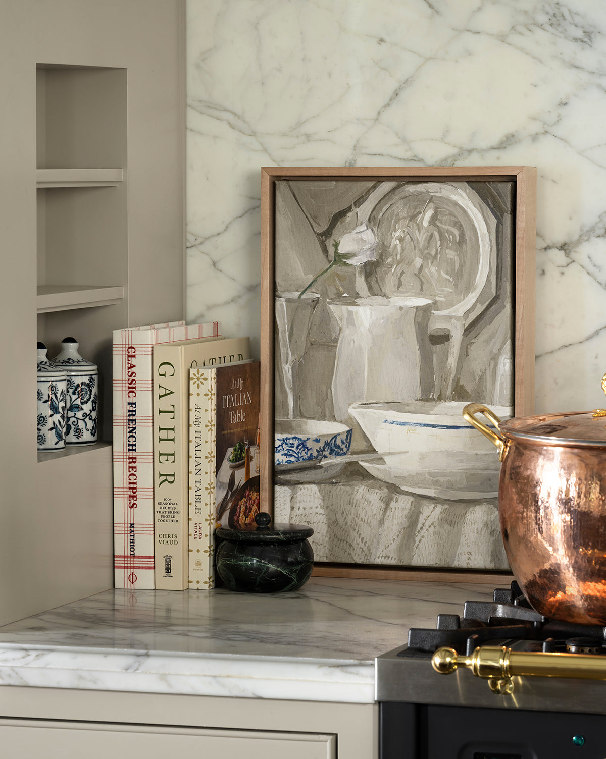 A kitchen countertop displays the "Classic French Recipes" cookbook, a decorative jar, marble mortar and pestle, framed still-life art, and a copper pot on the stove, all set against a marble backsplash, by Vendixs