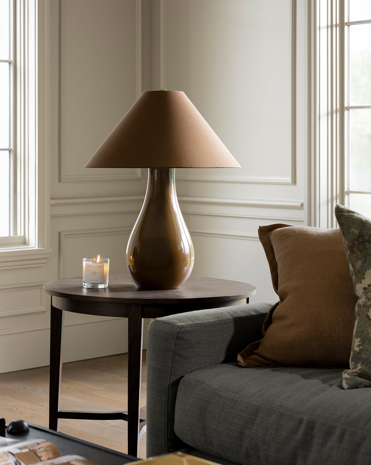 The Irving Table Lamp with a sculptural base and fabric shade sits beside a lit candle on a dark round side table next to a gray sofa with brown and patterned pillows, in an elegant room with tall windows and white panel walls, by Vendixs