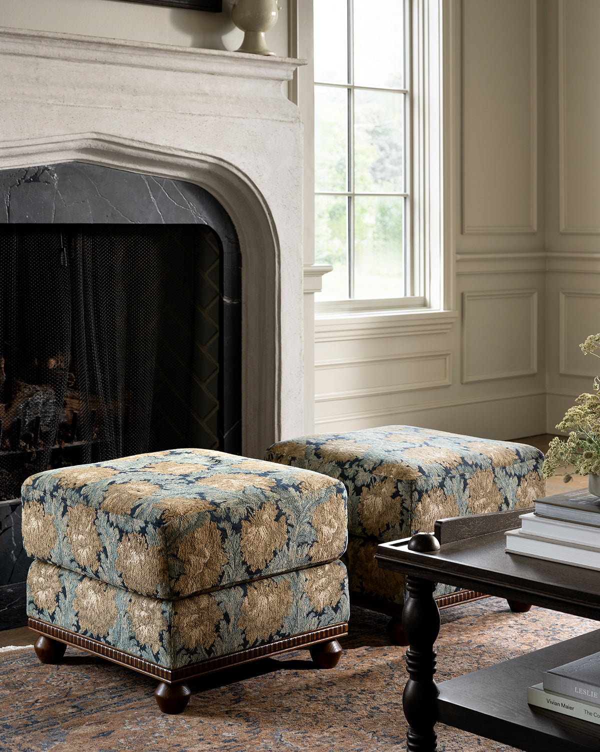 Two Catherine Ottomans, featuring a floral pattern and carved wood base, are placed in front of a fireplace with black marble surround in a bright living room with a window, coffee table, and beige walls, by Vendixs