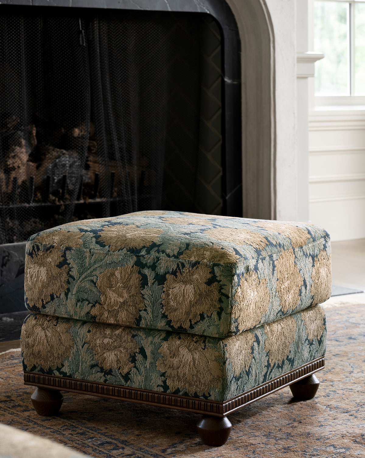 The Catherine Ottoman, featuring a carved wood base and floral upholstery, sits on a rug in front of a fireplace with a black screen in a bright, elegant living room, by Vendixs