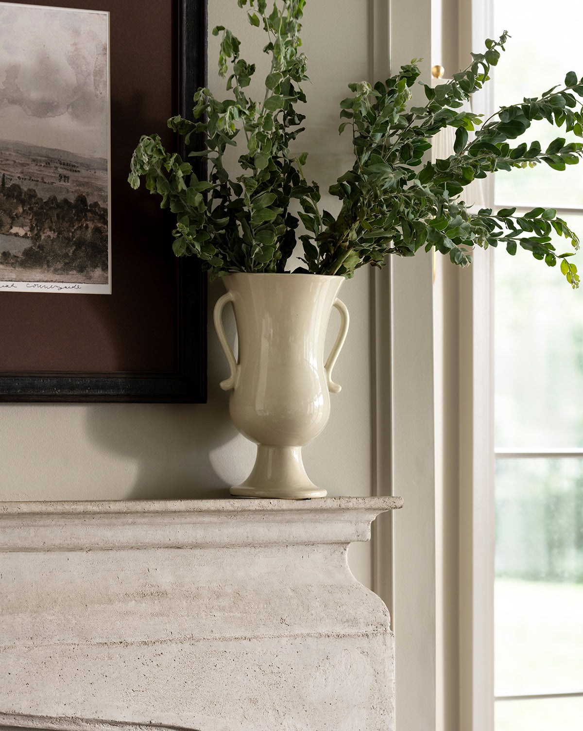 The Delphine Footed Vase, cream-colored and elegant with two handles, holds leafy green branches on a stone mantel beside a framed picture and a sunlit window, by McGee & Co.