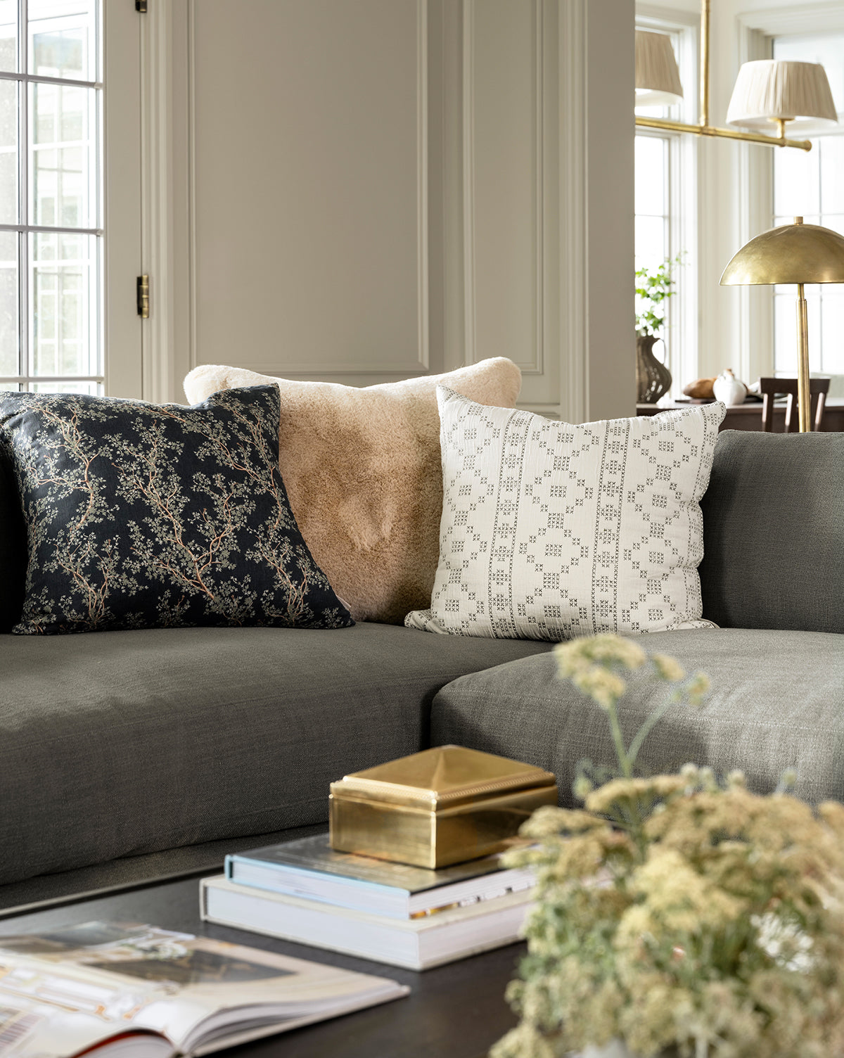 A gray sofa with three decorative pillows, including a botanical one and the Antigone Pillow Cover, sits by large windows. In front is a coffee table with stacked books, a gold box, and a vase of dried flowers by Vendixs